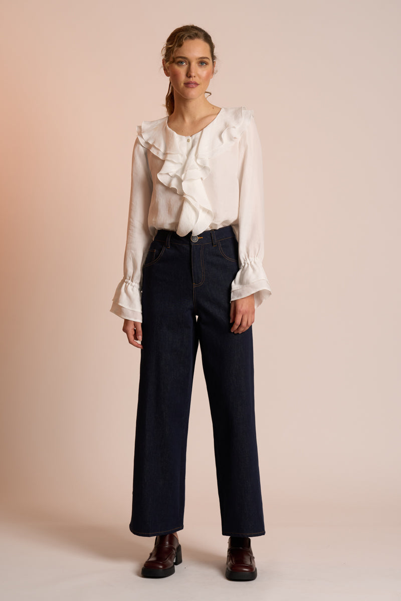 Woman wearing a white blouse with ruffled details and dark pants on a beige background