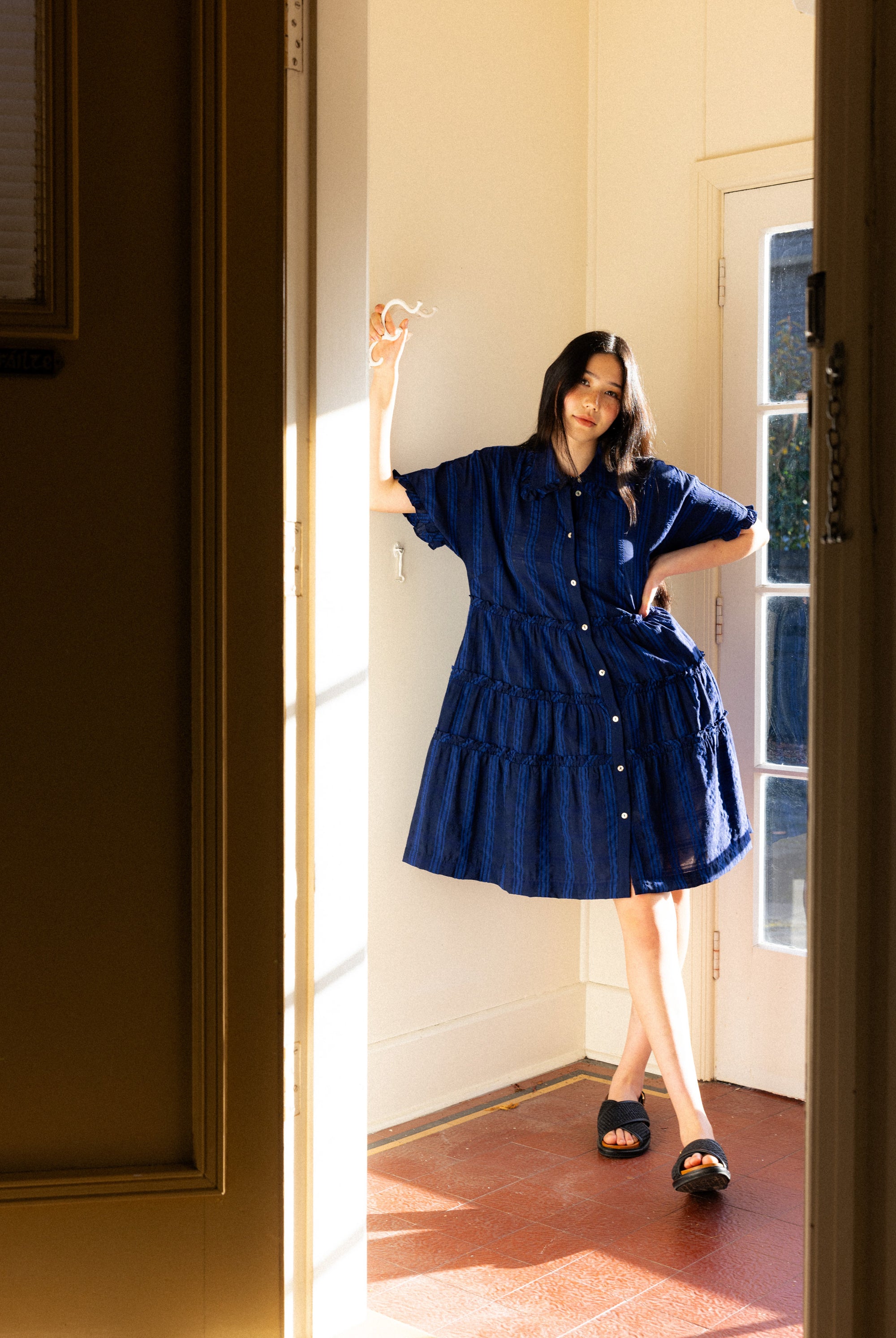 Woman in a blue dress standing in a sunlit room with a door open.