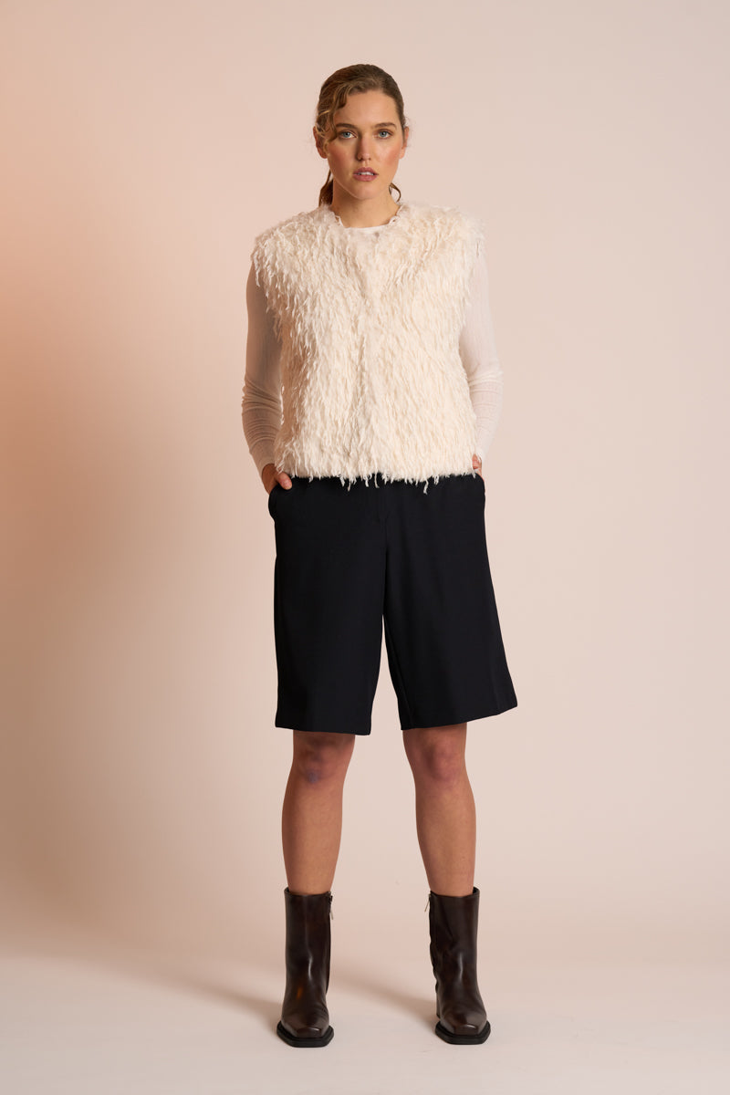 Woman wearing an off white gilet, black shorts, and brown boots on a beige background