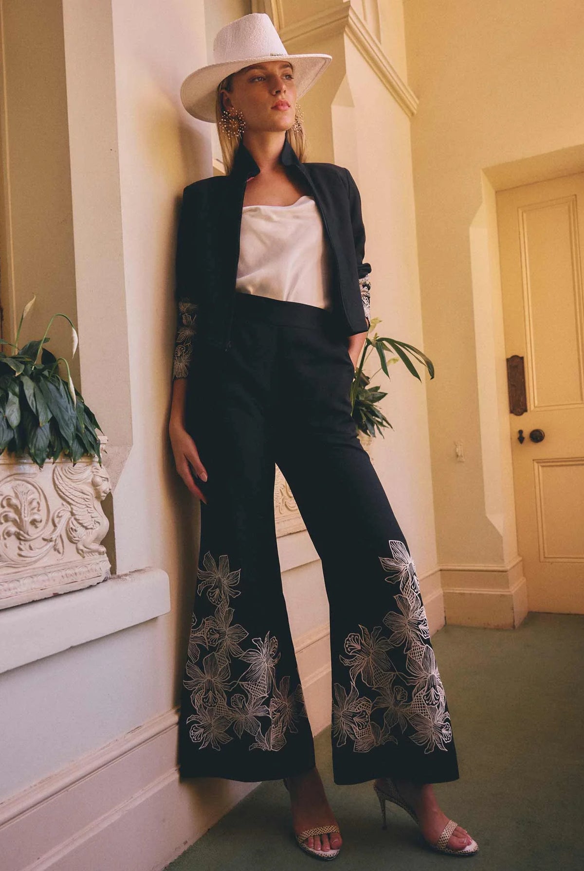 women wearing a pair of wide leg black pants with floral embroidery and a matching black jacket with a white hat