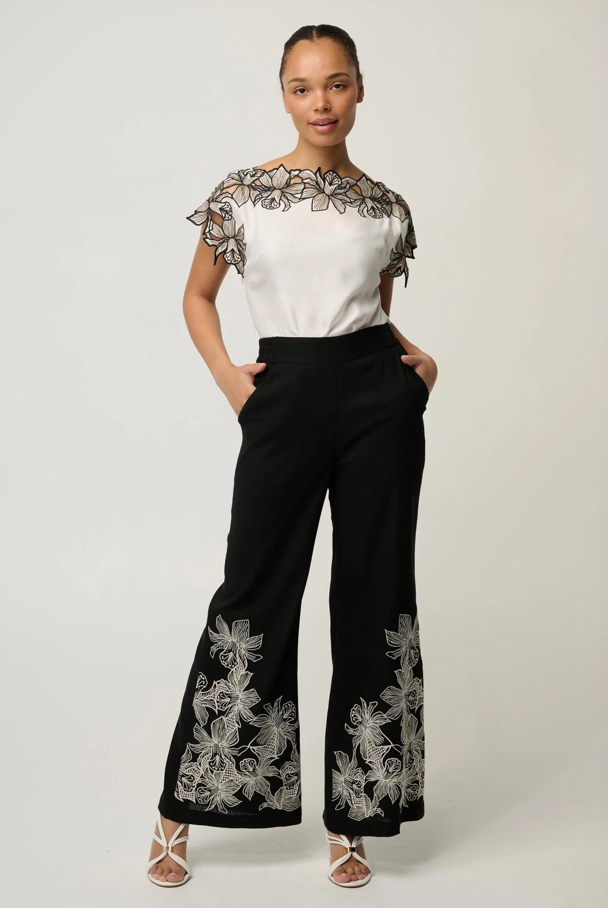women wearing a white top with black floral embroidery and black pants with with white embroidery on a plain background