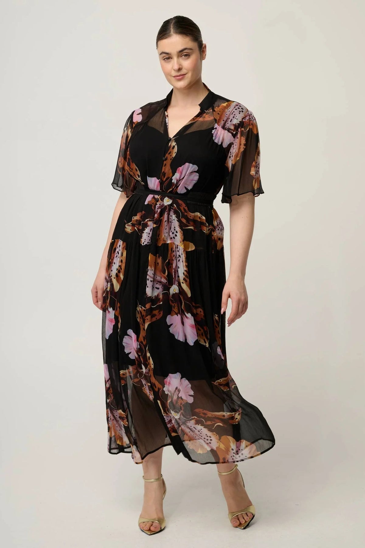 Woman wearing a black floral floaty chiffon dress with tie waist and short sleeves and gold sandals against a white background