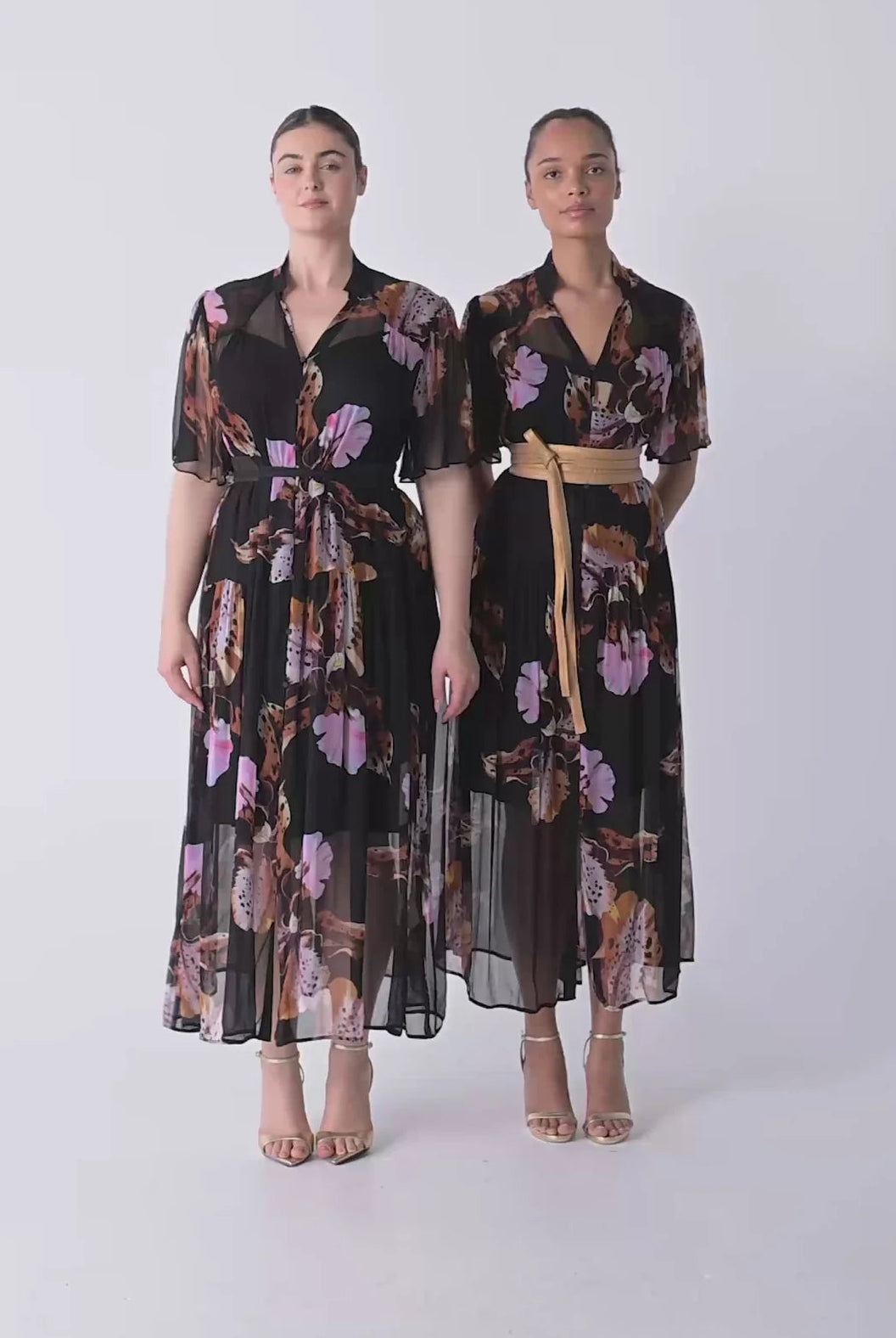 Woman wearing a black floral floaty chiffon dress with tie waist and short sleeves and gold sandals against a white background