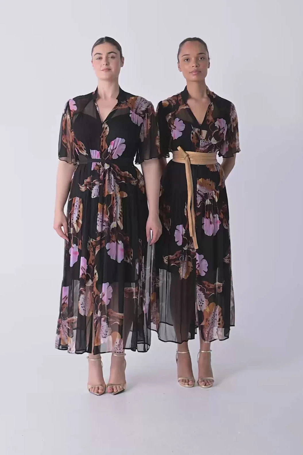 Woman wearing a black floral floaty chiffon dress with tie waist and short sleeves and gold sandals against a white background