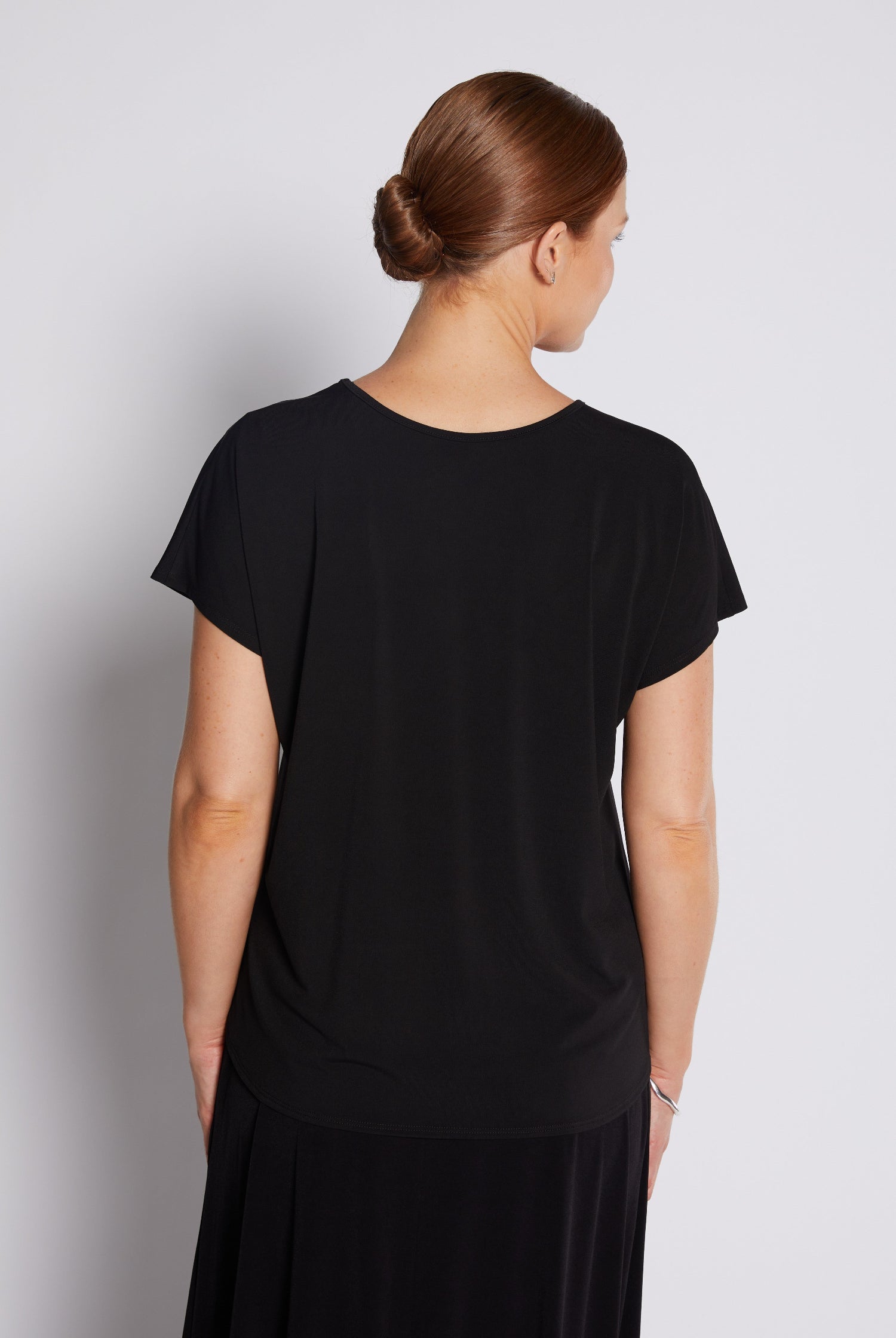 A woman viewed from the back wearing a black short-sleeve top and a black skirt against a light gray studio background.