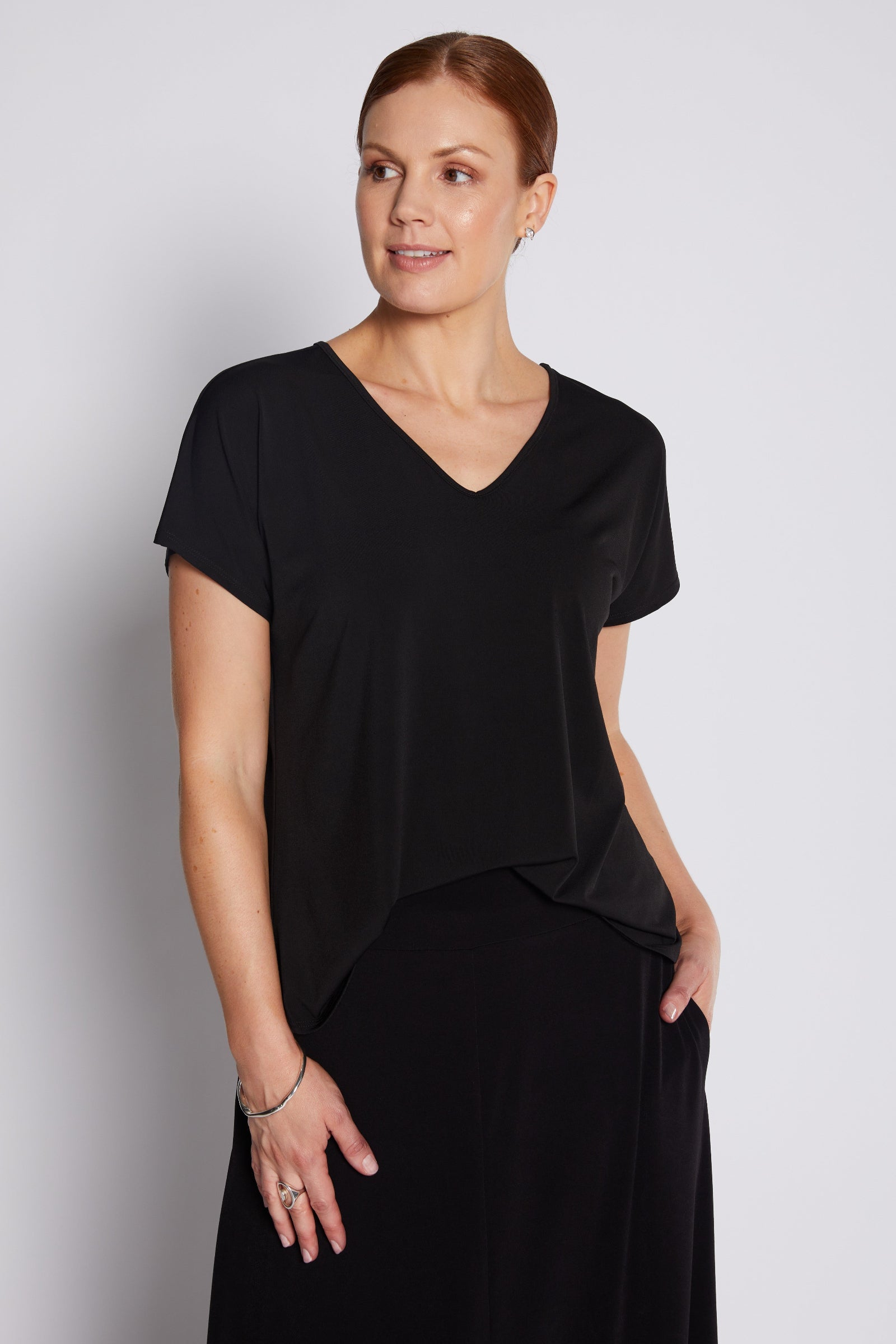 A woman modeling a black short-sleeve V-neck top with an asymmetrical draped hem, paired with a black skirt, against a light gray studio background.