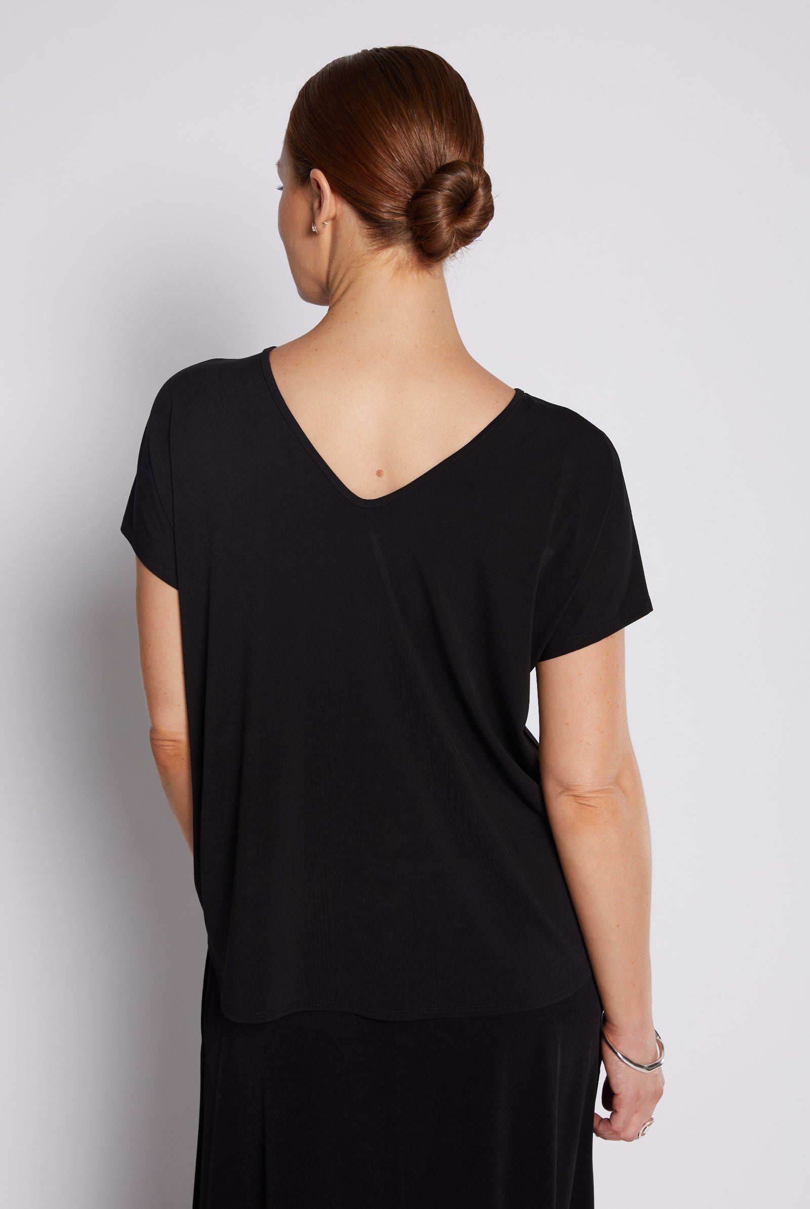 A woman viewed from the back wearing a black short-sleeve V-neck top and black skirt against a light gray studio background.