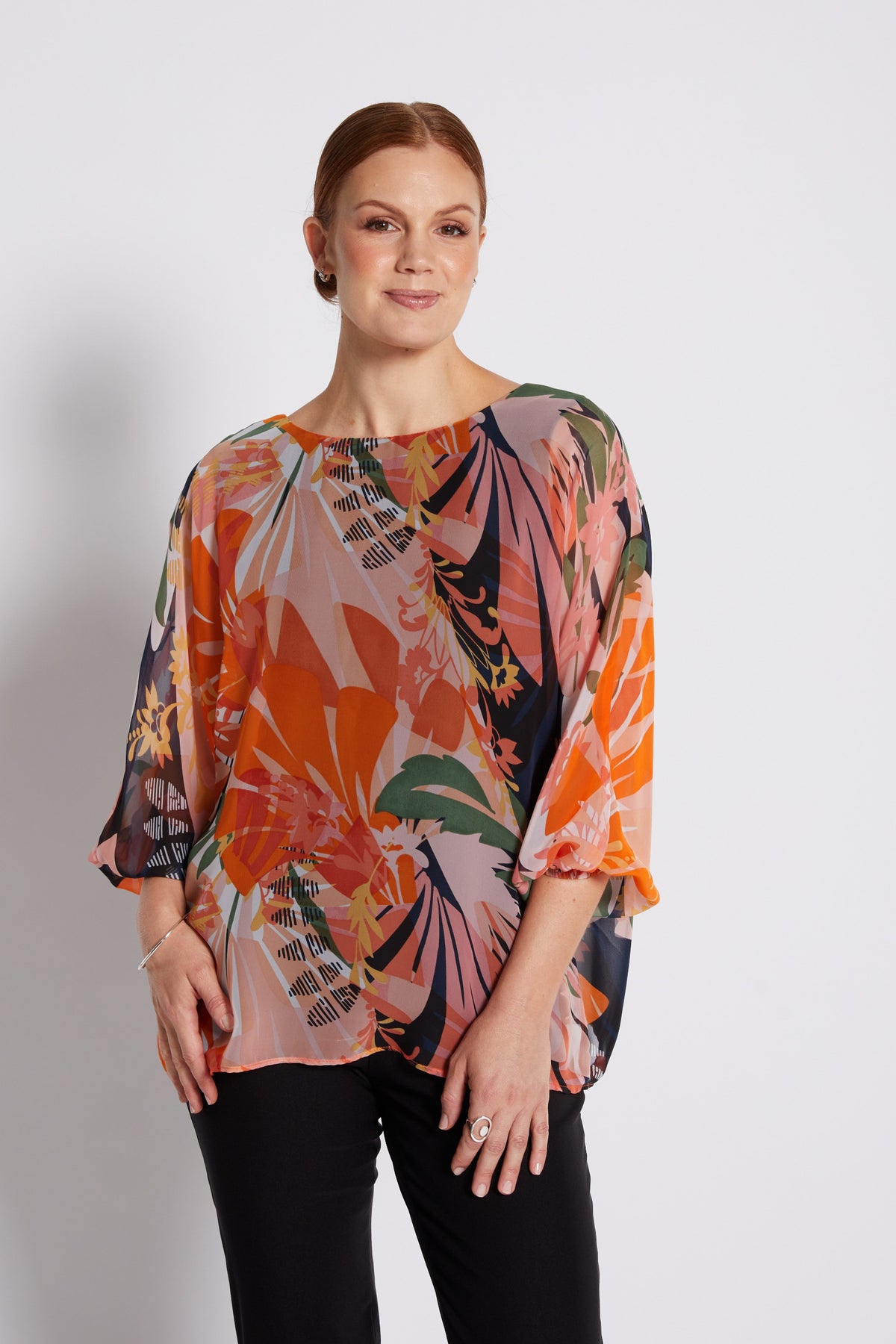 A woman modeling a loose, short-sleeved blouse with a vibrant multicolor tropical floral print in orange, pink, green, and black, paired with black pants, against a plain light gray background.