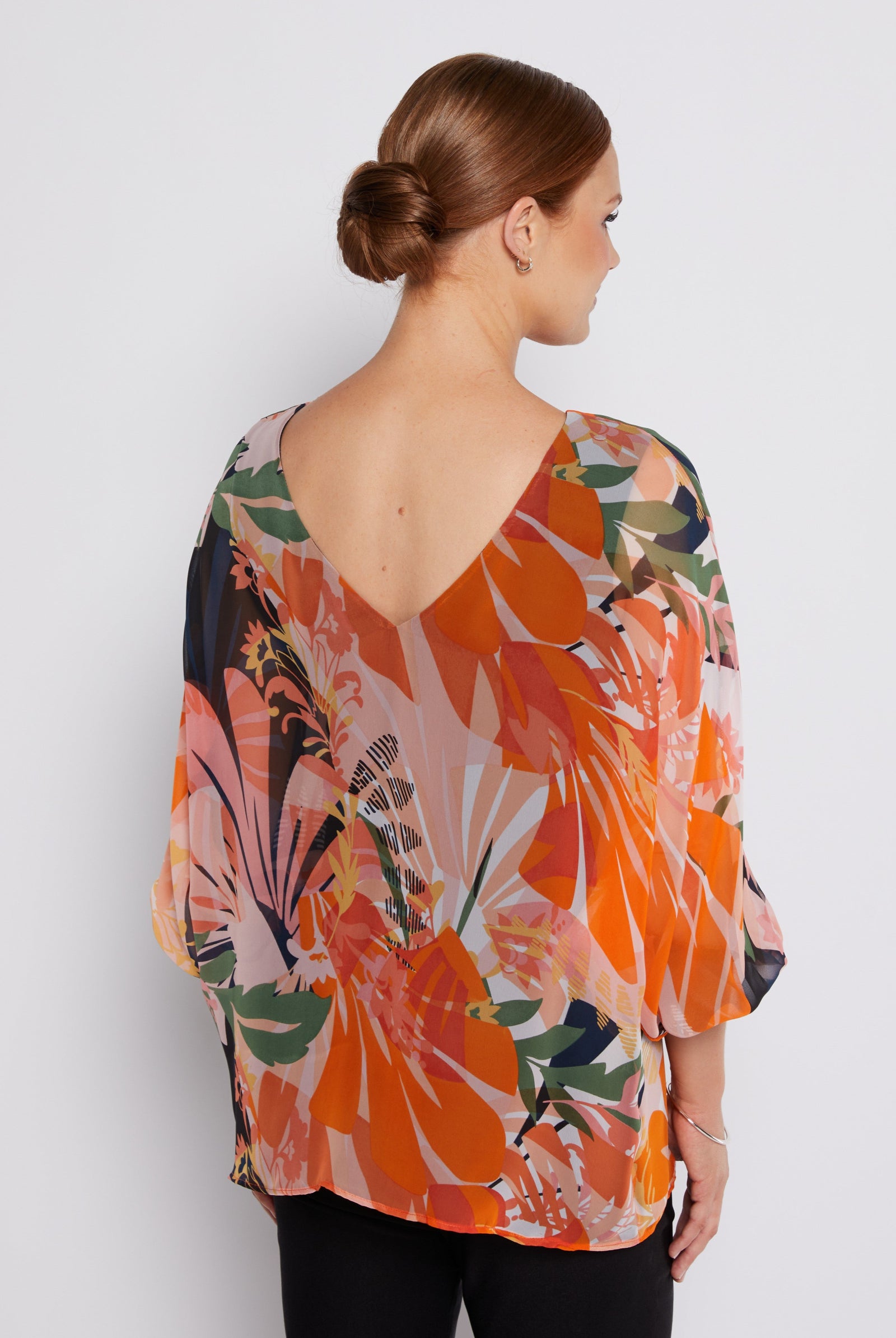 A woman shown from the back wearing a loose, V-neck blouse with a bold tropical floral print in orange, pink, green, and beige, paired with black pants against a light gray studio background.