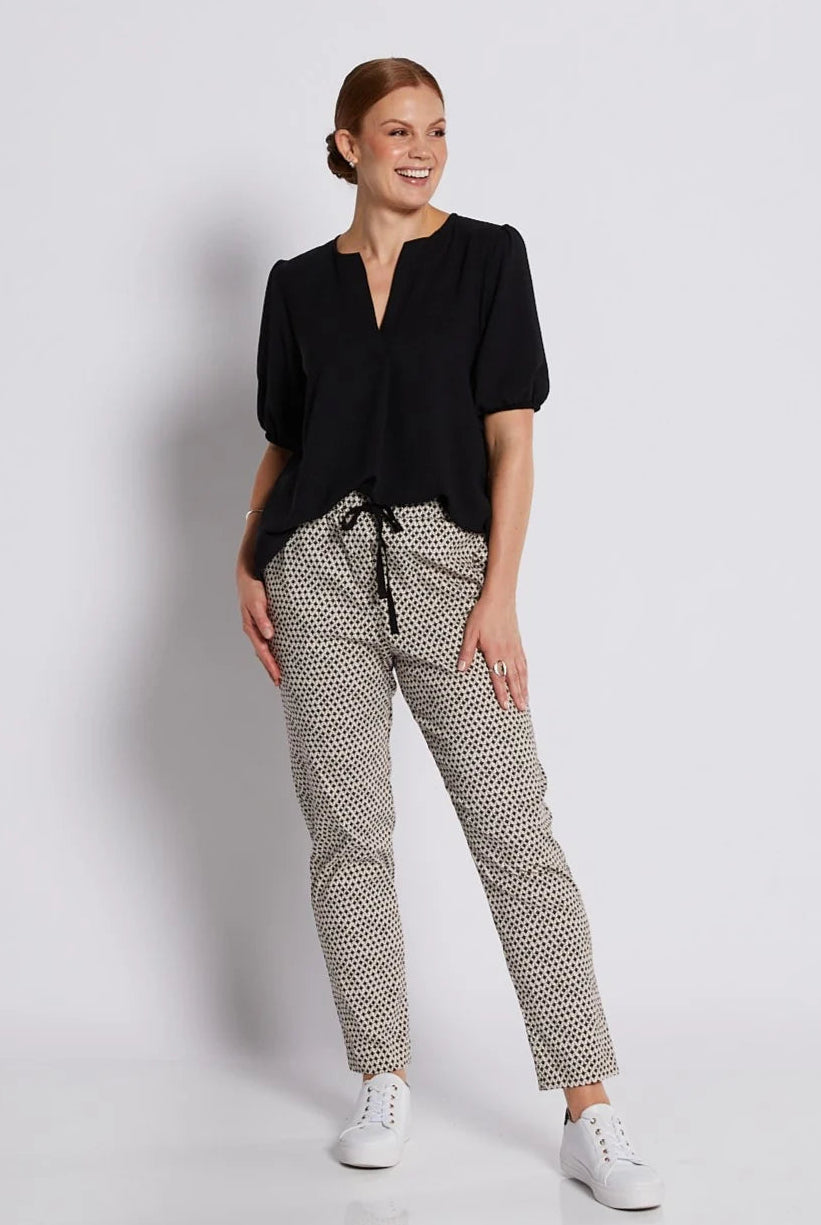 A woman modeling a black short-sleeve blouse and beige-and-black patterned trousers with white sneakers, standing against a light gray studio background.