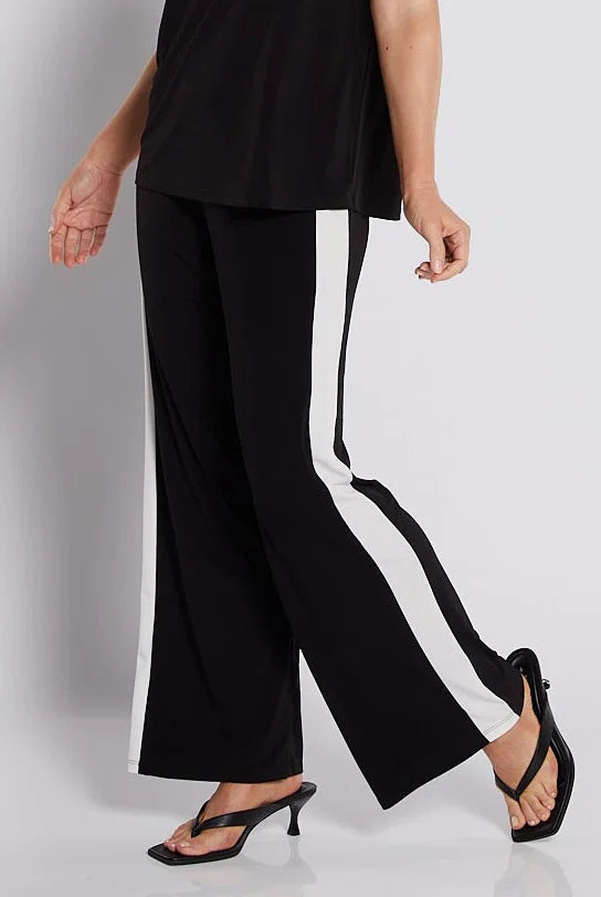 A woman modeling a black two-piece set consisting of a short-sleeve top with white shoulder stripes and matching wide-leg trousers with white side stripes, paired with black strappy heels, against a light gray studio background.