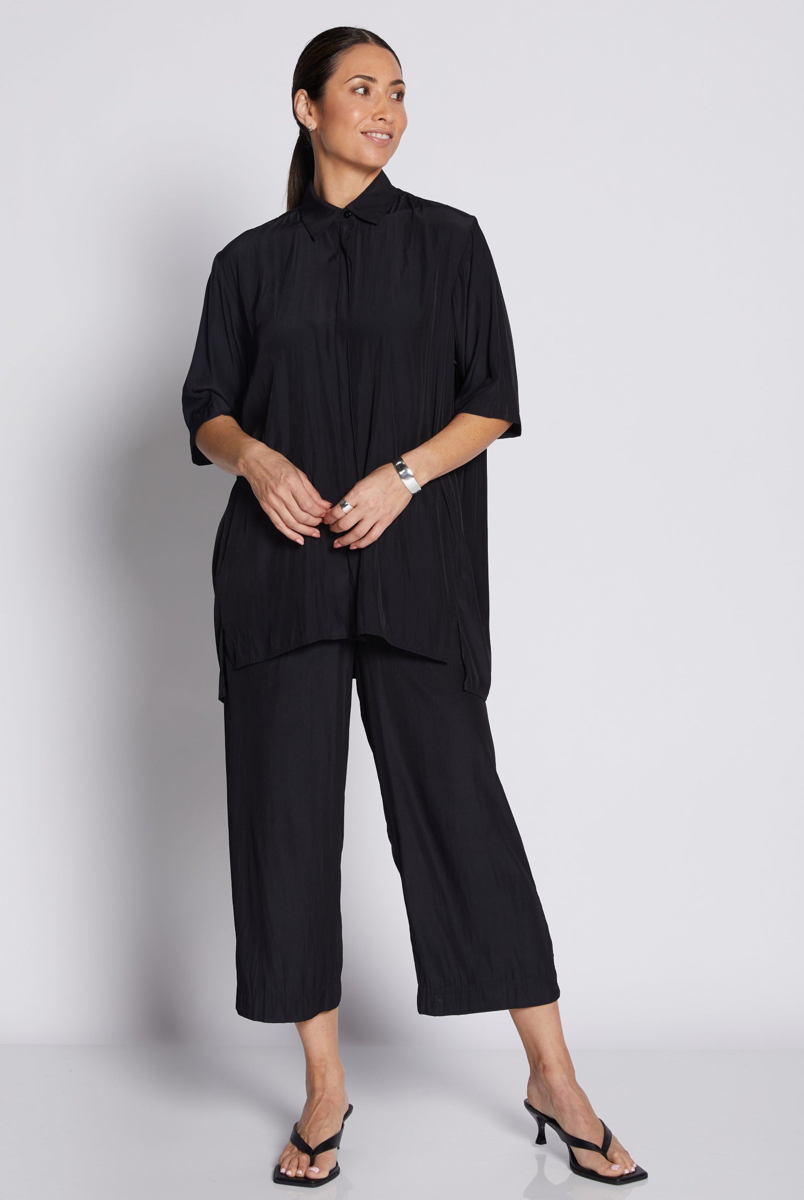 A woman modeling a black two-piece outfit consisting of a short-sleeve, high-neck, pleated blouse and matching cropped pants, paired with black sandals against a light gray studio background.