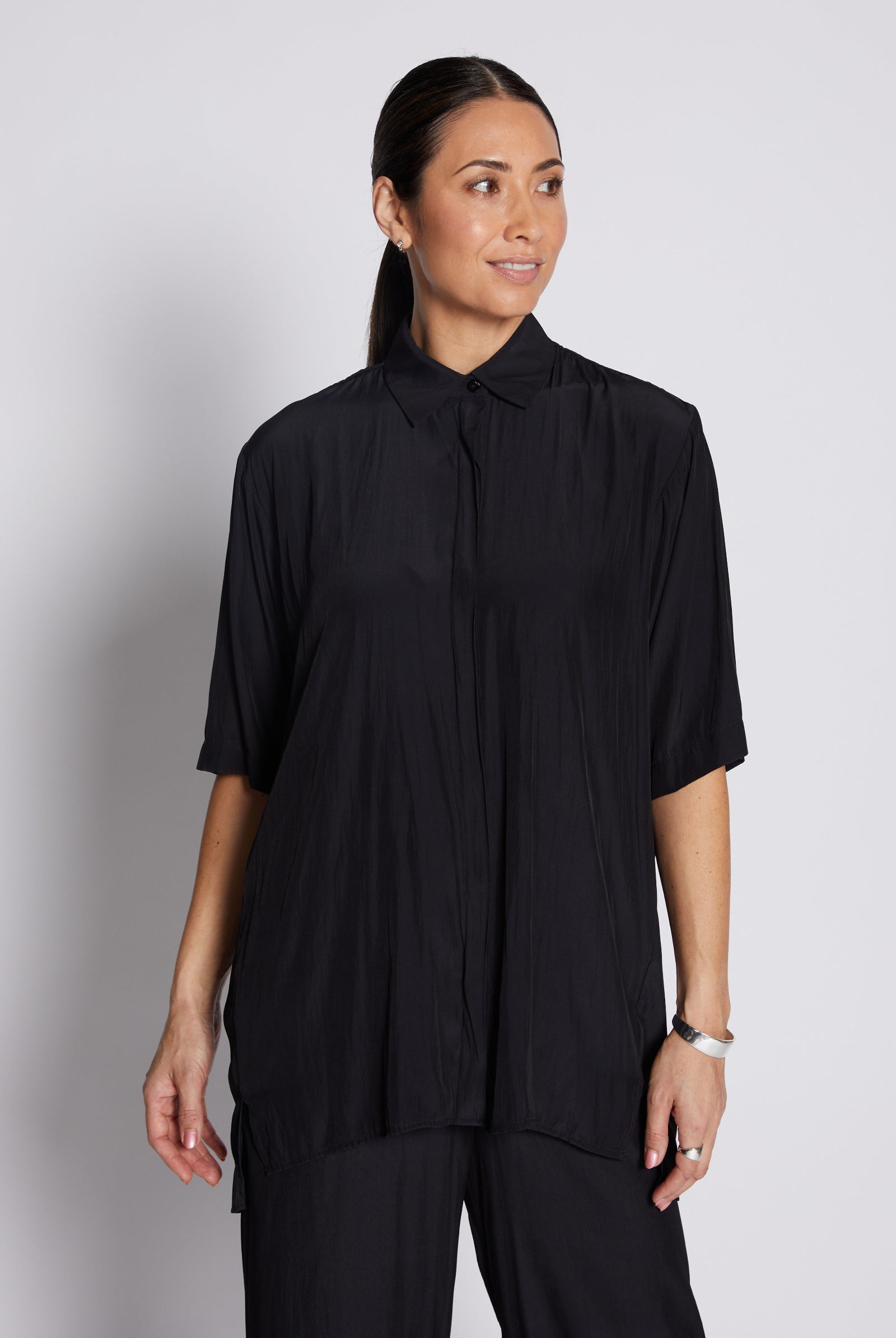 A woman modeling a black short-sleeve button-up shirt with a high collar and subtle vertical pleating, paired with black trousers, against a light gray studio background.