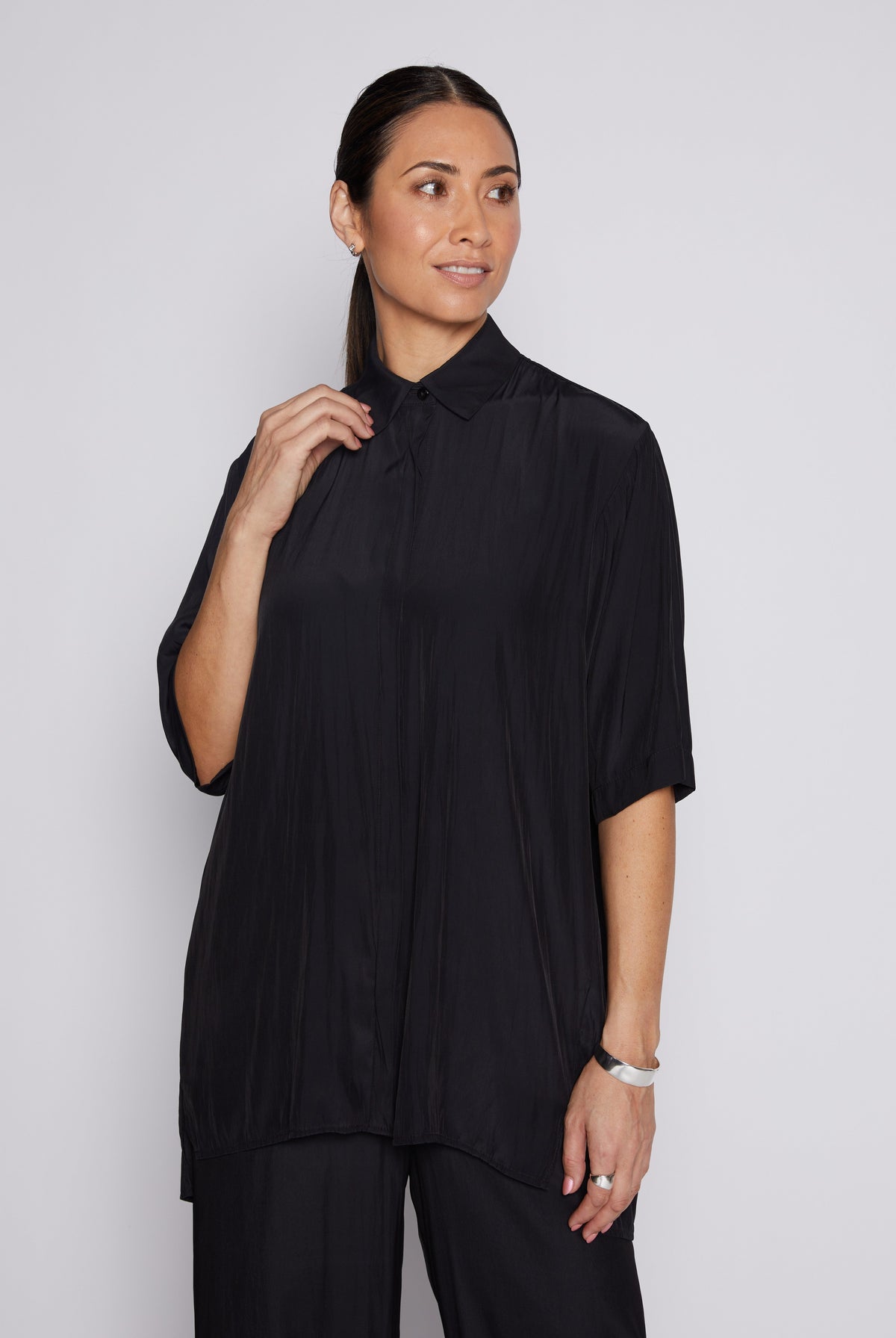 A woman modeling a black short-sleeve pleated button-up shirt with a collar, paired with black trousers, against a light gray studio background.