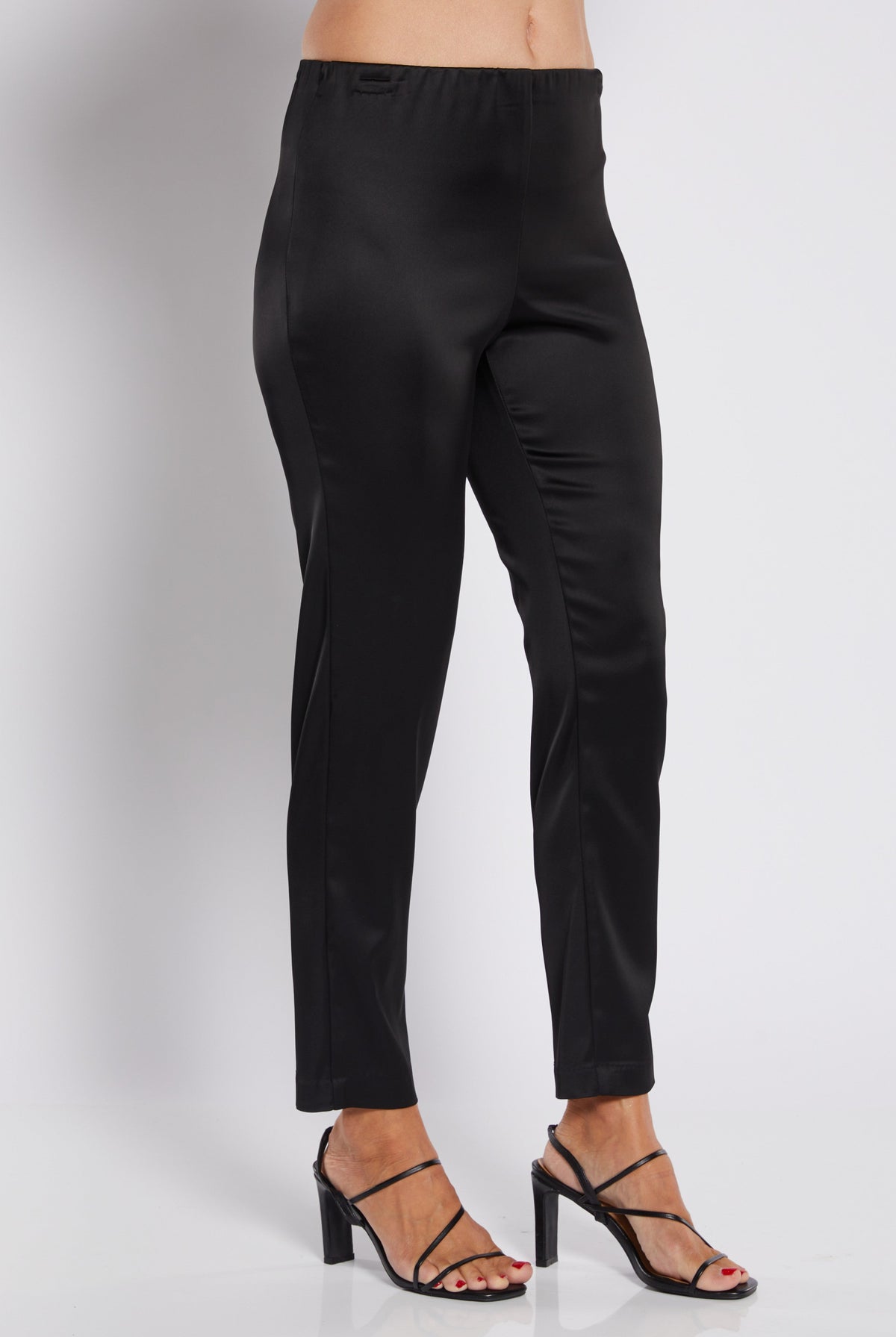 Black slim-fit ankle-length pants shown on a model from the waist down, paired with black strappy high-heeled sandals against a light gray studio background.