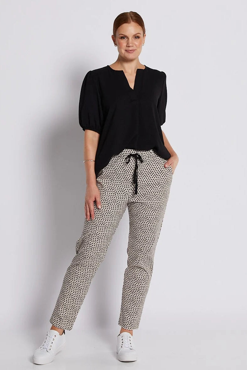 A woman modeling a black short-sleeve top with a split neckline and patterned trousers featuring a small black-on-beige print, paired with white sneakers, against a light gray studio background.