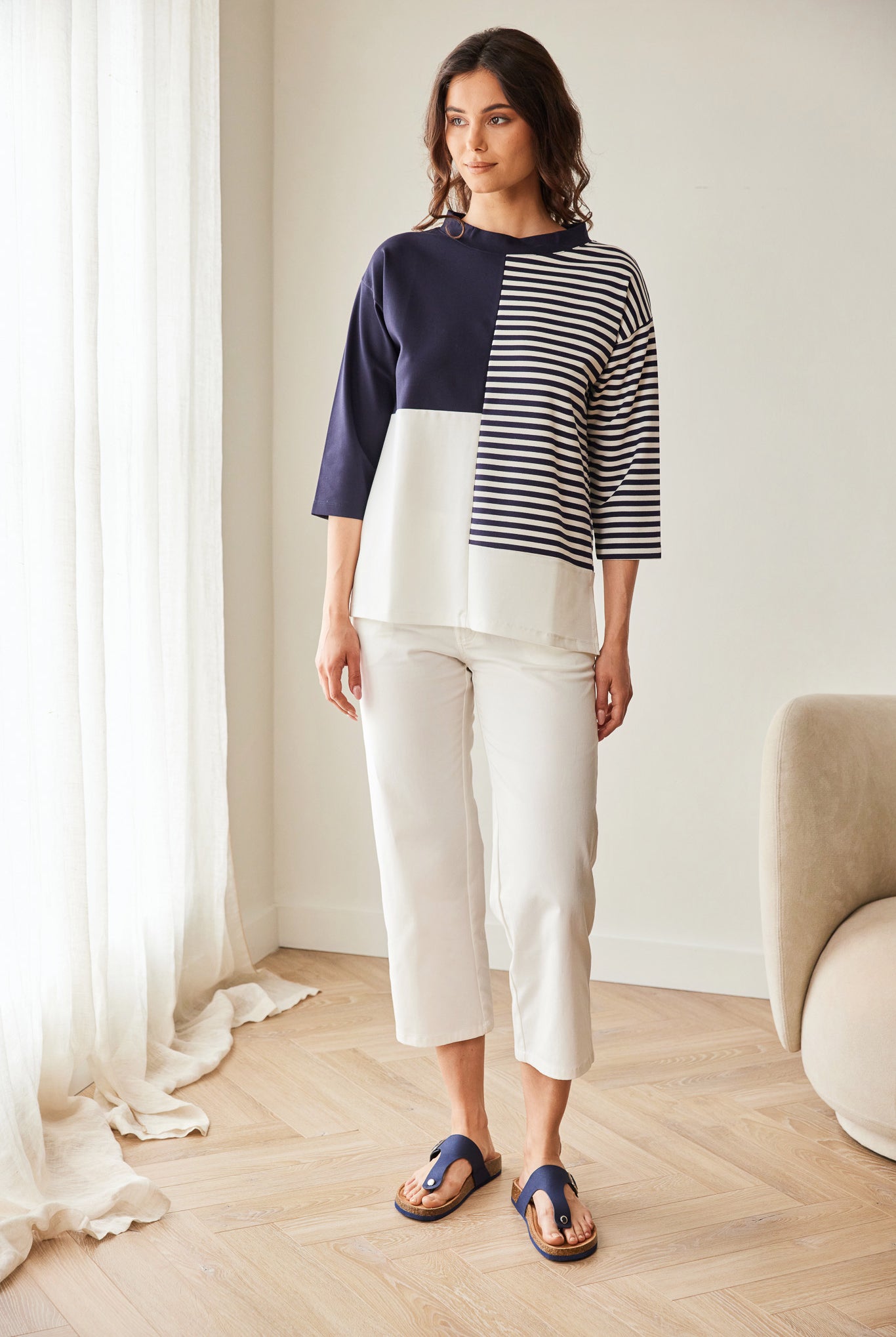 Woman wearing a color-blocked sweater and white pants in a room with light wood flooring and a white wall.
