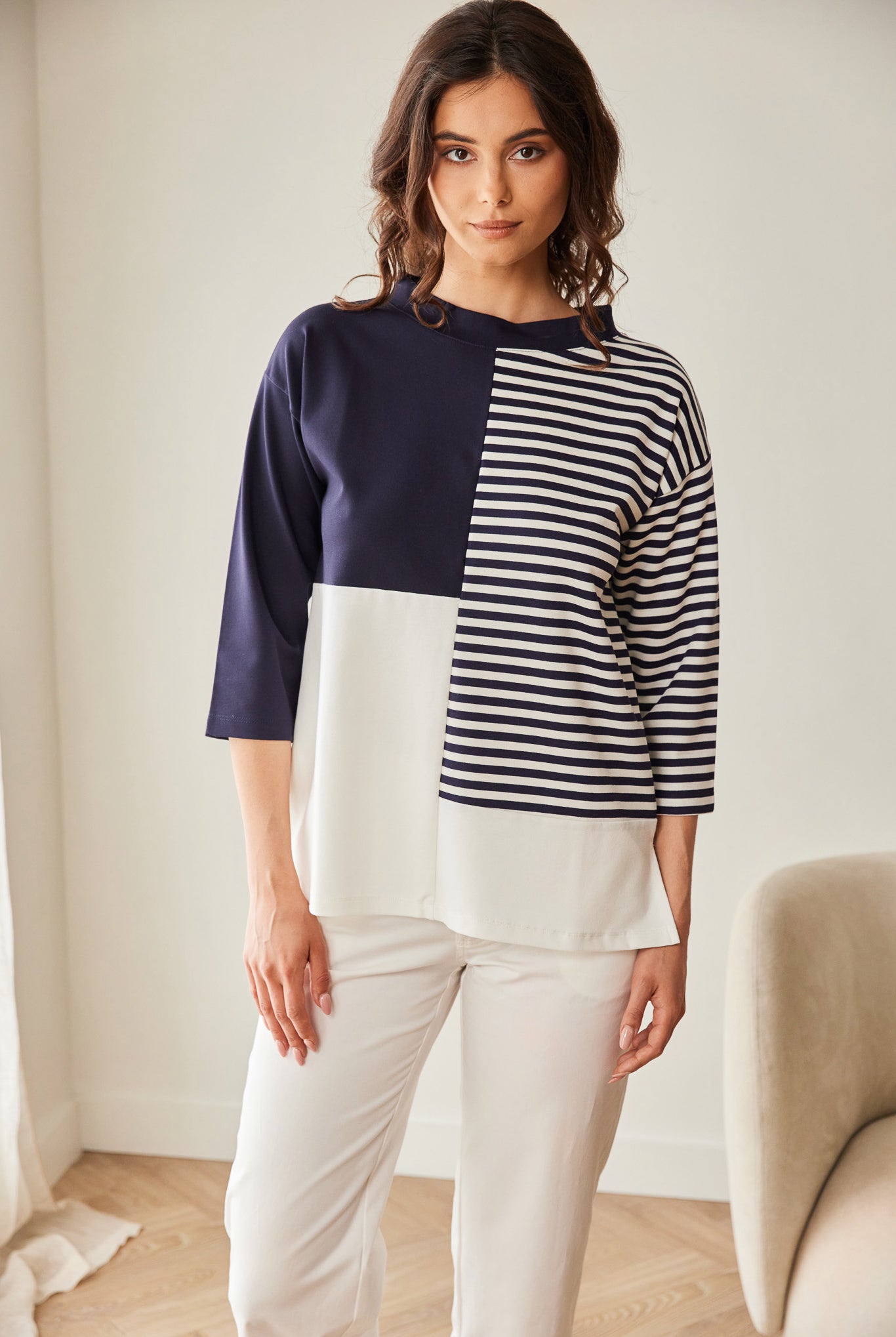 Woman wearing a color-blocked sweater and white pants in a minimalistic room.