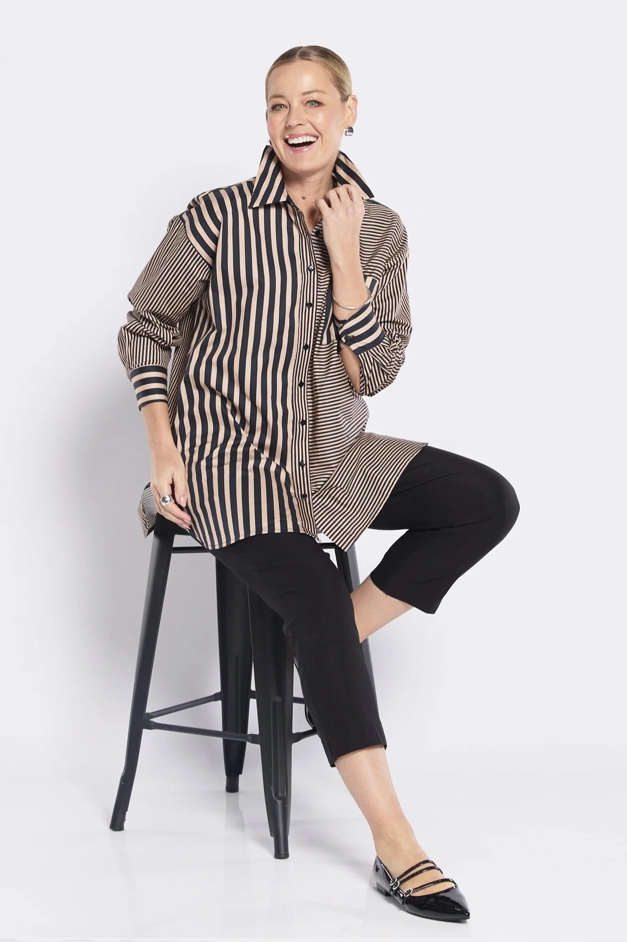 Woman sitting on a stool wearing a striped shirt and black pants against a white background