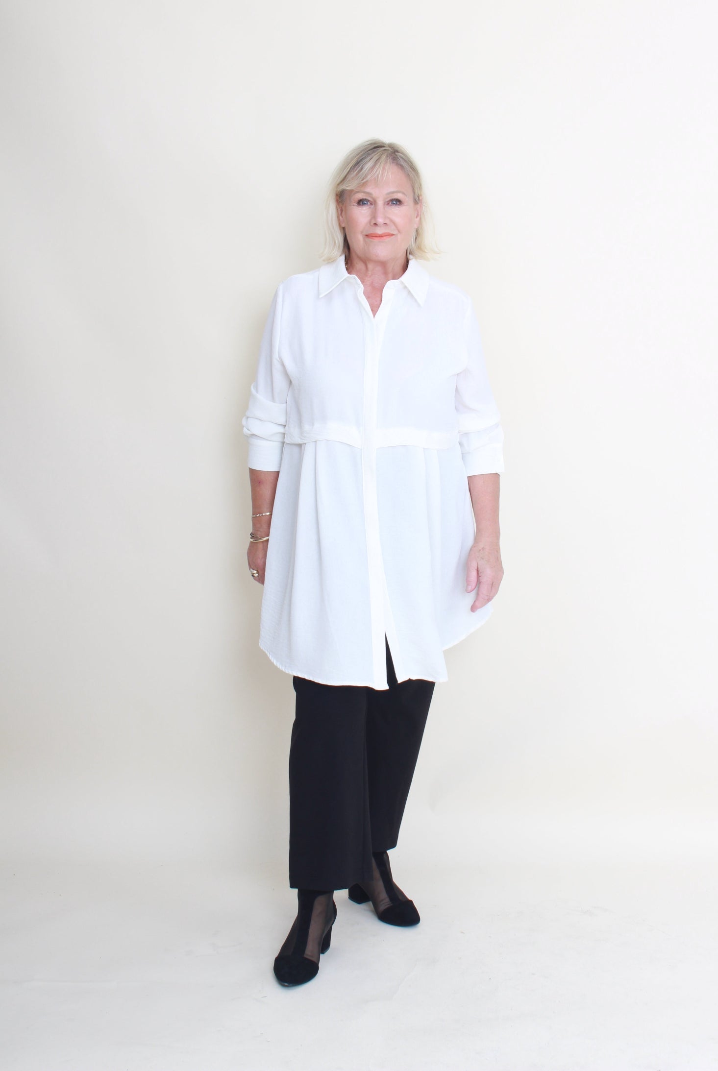 An older woman stands against a plain off-white studio background wearing a white long-sleeve button-up shirt with black wide-leg pants and black flats.