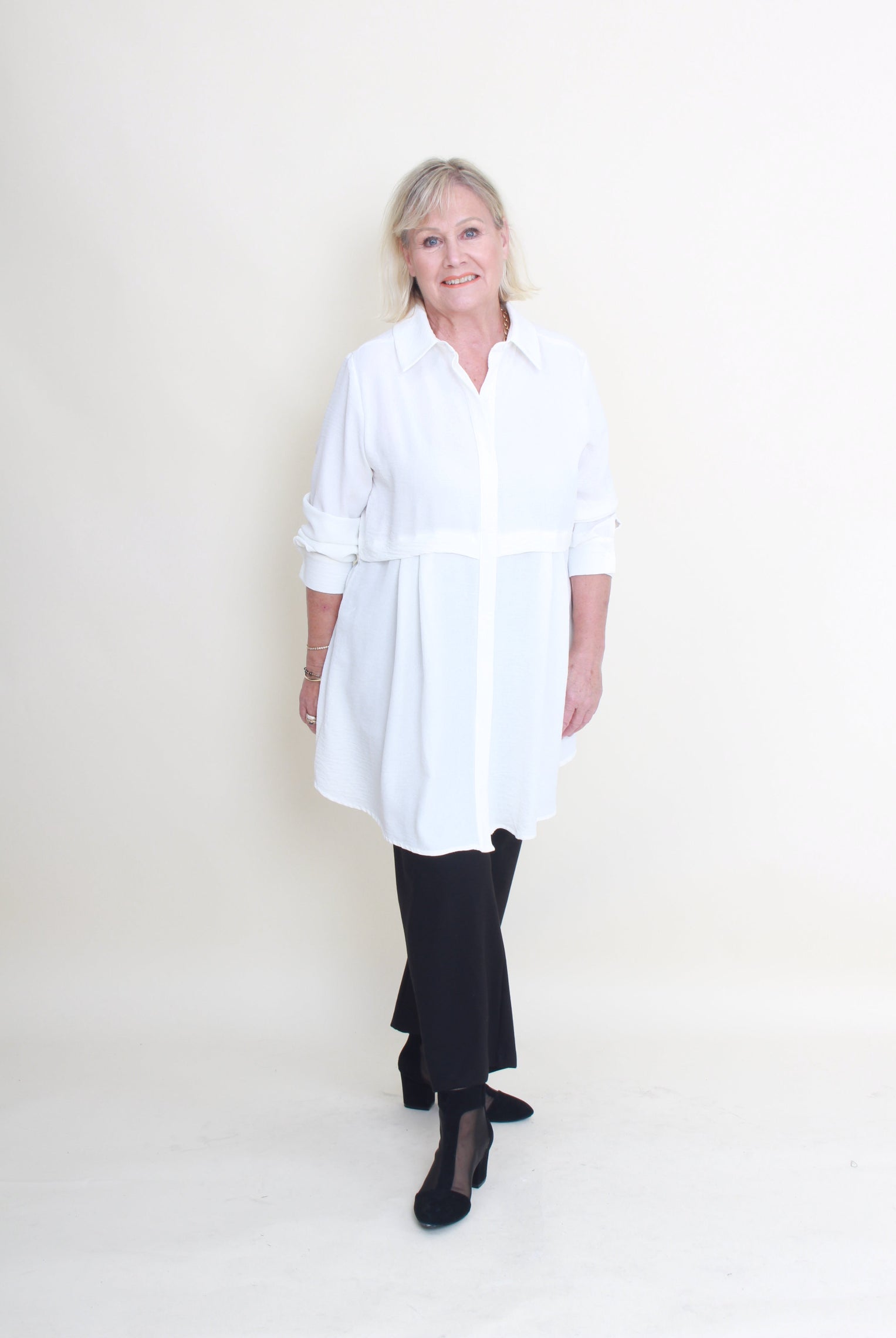 A woman modeling a white long-sleeve button-up shirt dress with a tie waist, paired with black pants and black heels, standing against a plain off-white studio background.