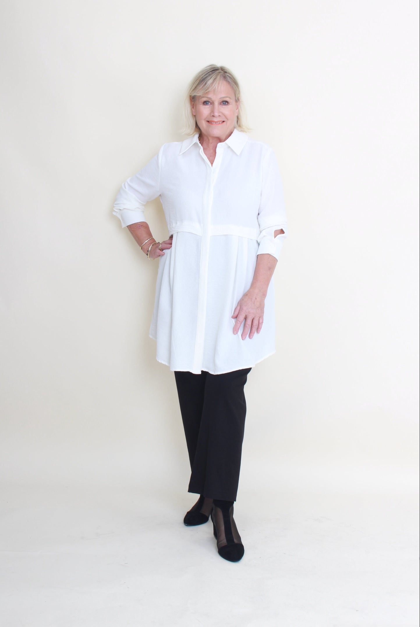 An older woman lying flat on a plain off-white studio floor, wearing a loose white long-sleeve top or dress over black pants and black shoes.