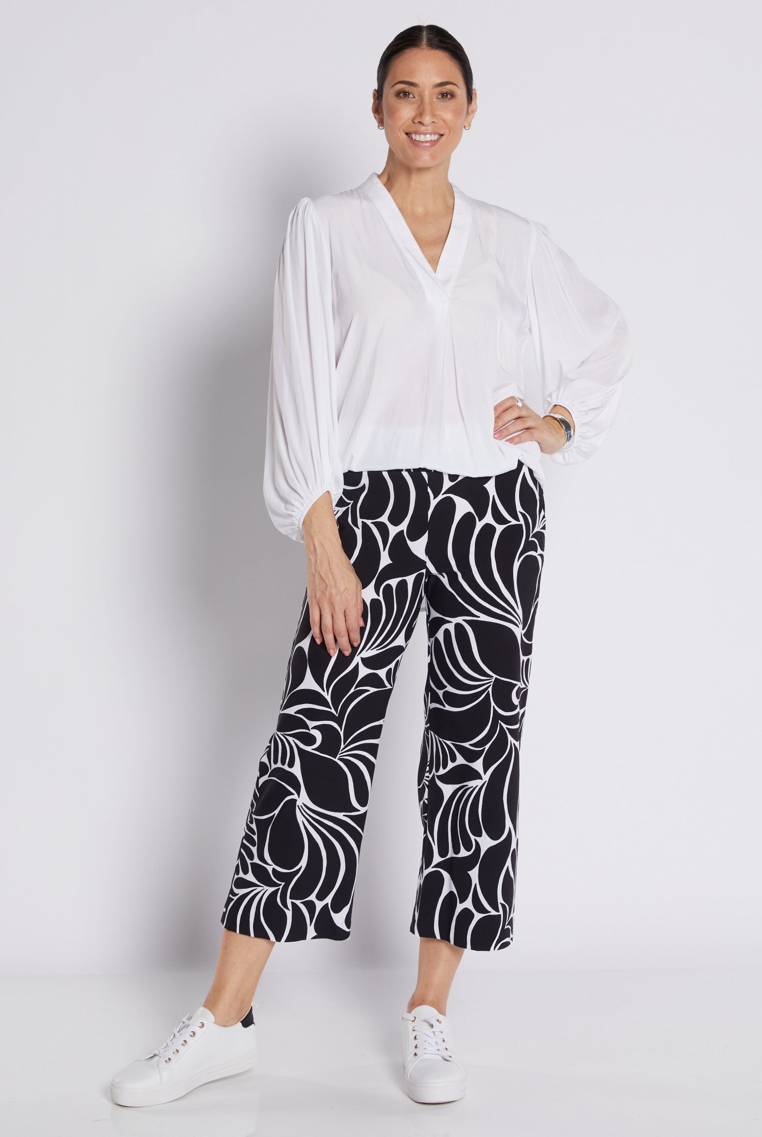 A woman modeling a white long-sleeve blouse and black cropped pants with a bold white floral pattern, paired with white sneakers, against a light gray studio background.