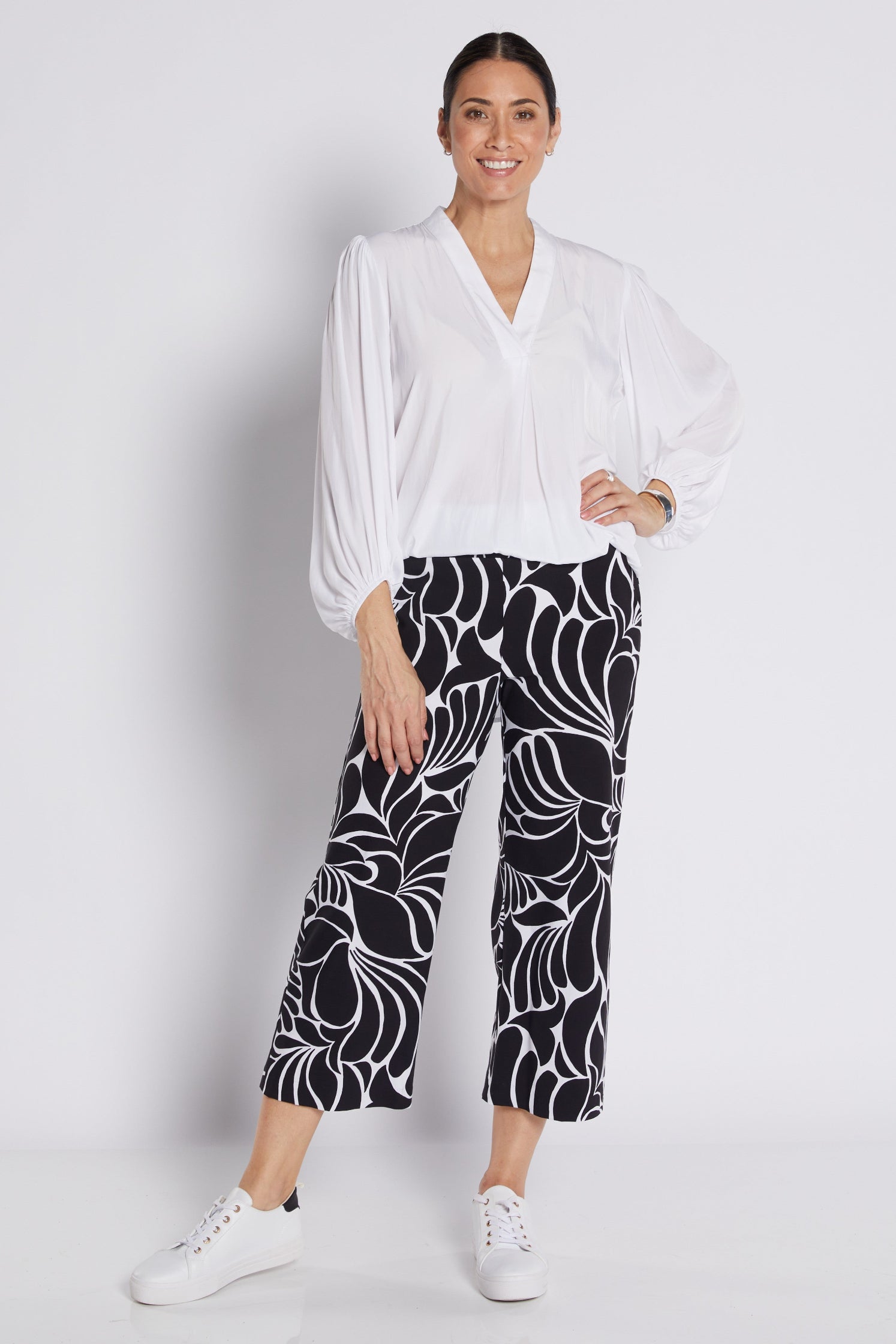 A woman modeling a white long-sleeve blouse and black cropped pants with a bold white floral pattern, paired with white sneakers, against a light gray studio background.