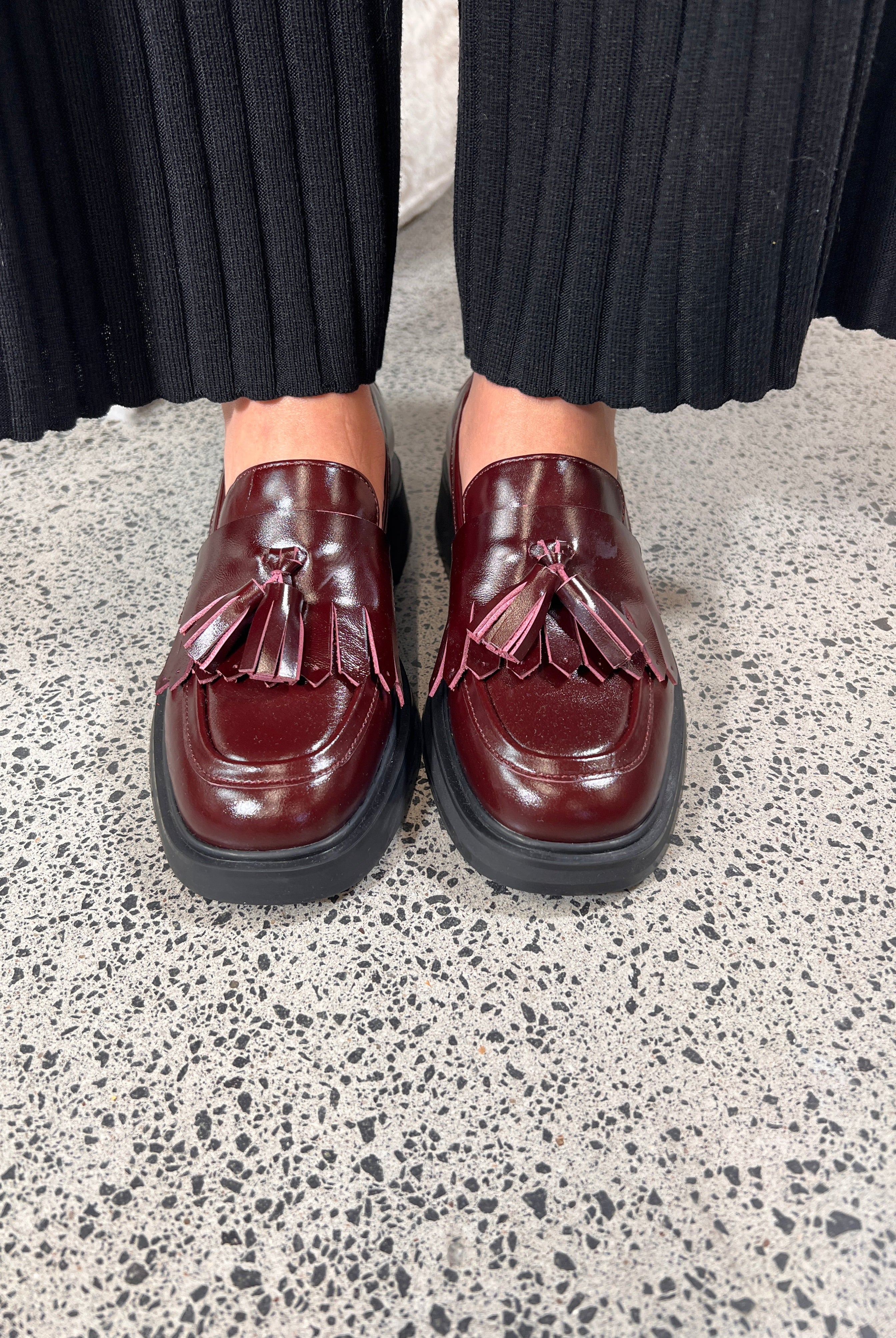 Person wearing maroon loafers with tassels on a speckled gray floor.