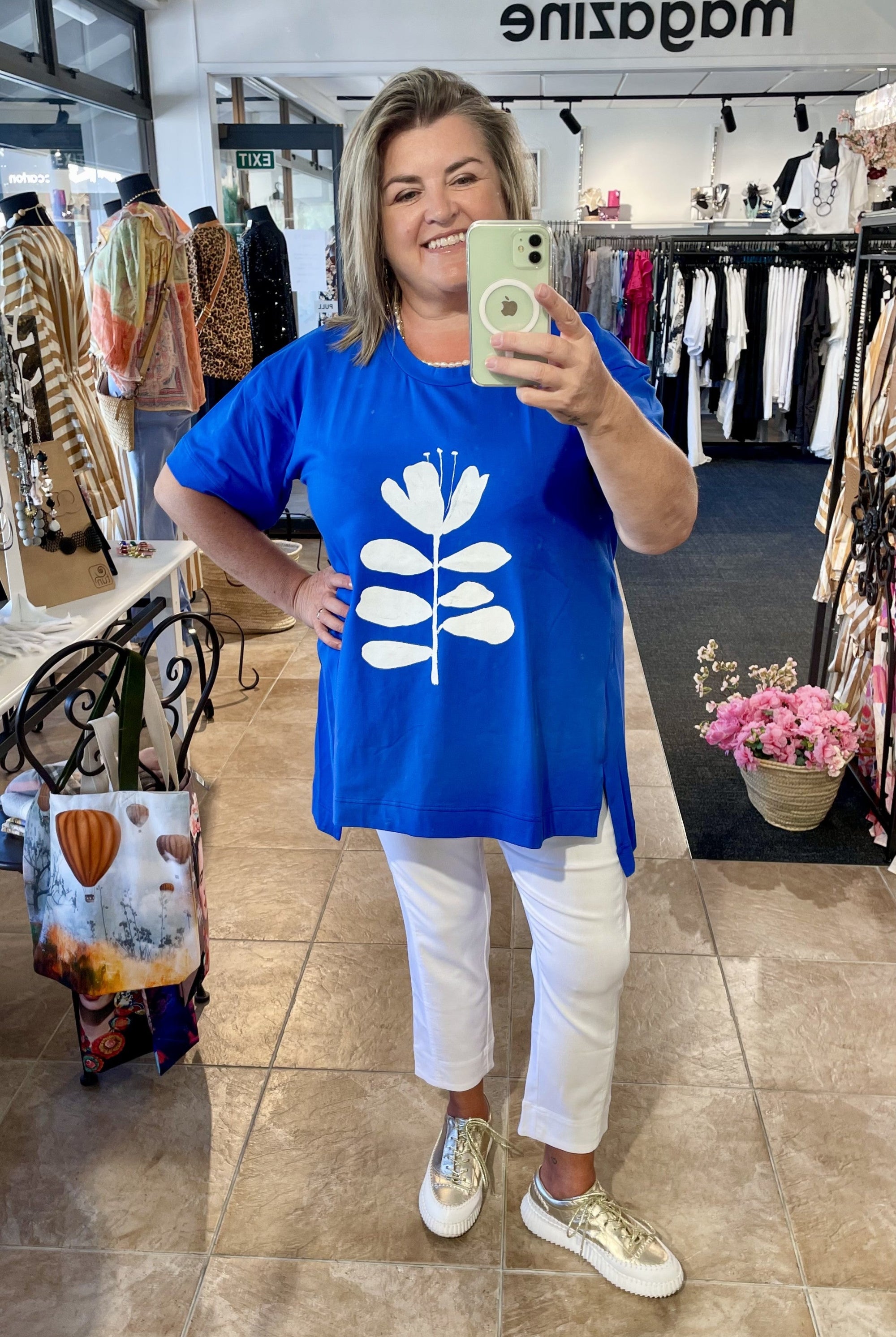 Woman taking a mirror selfie wearing a blue t-shirt with a white floral design in a clothing store.
