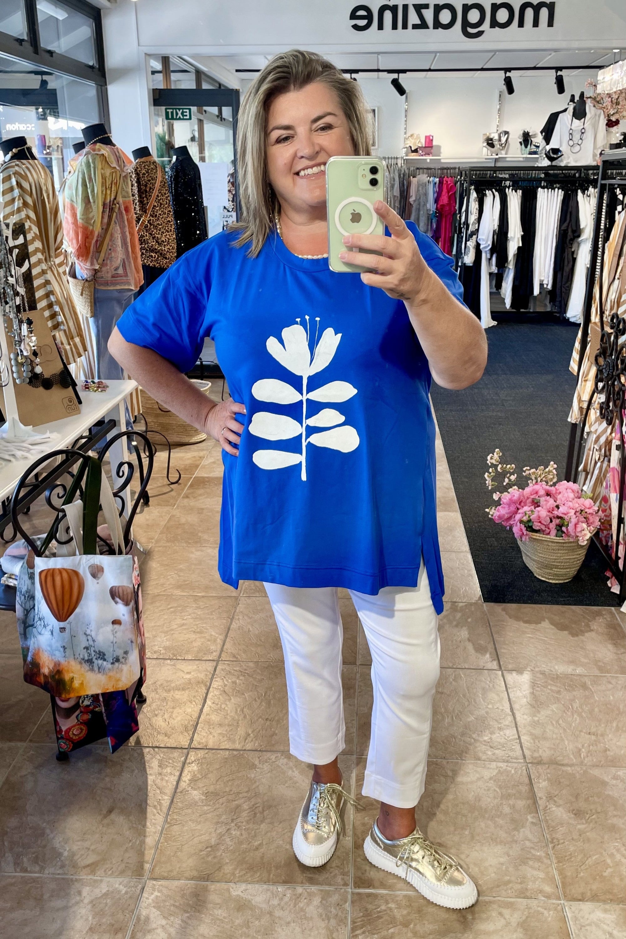 Woman taking a mirror selfie wearing a blue t-shirt with a white floral design in a clothing store.