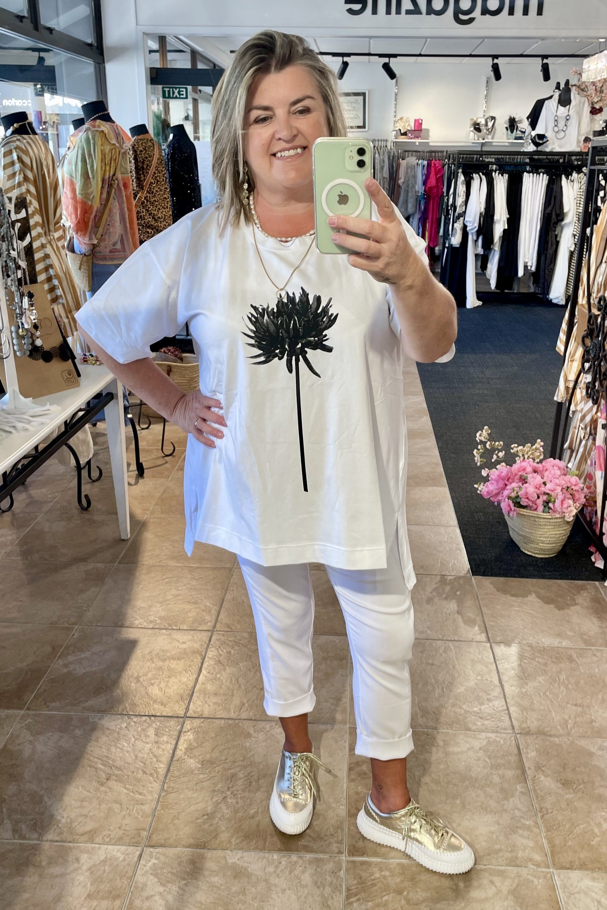 Woman taking a mirror selfie in a clothing store, wearing a white outfit with gold shoes.