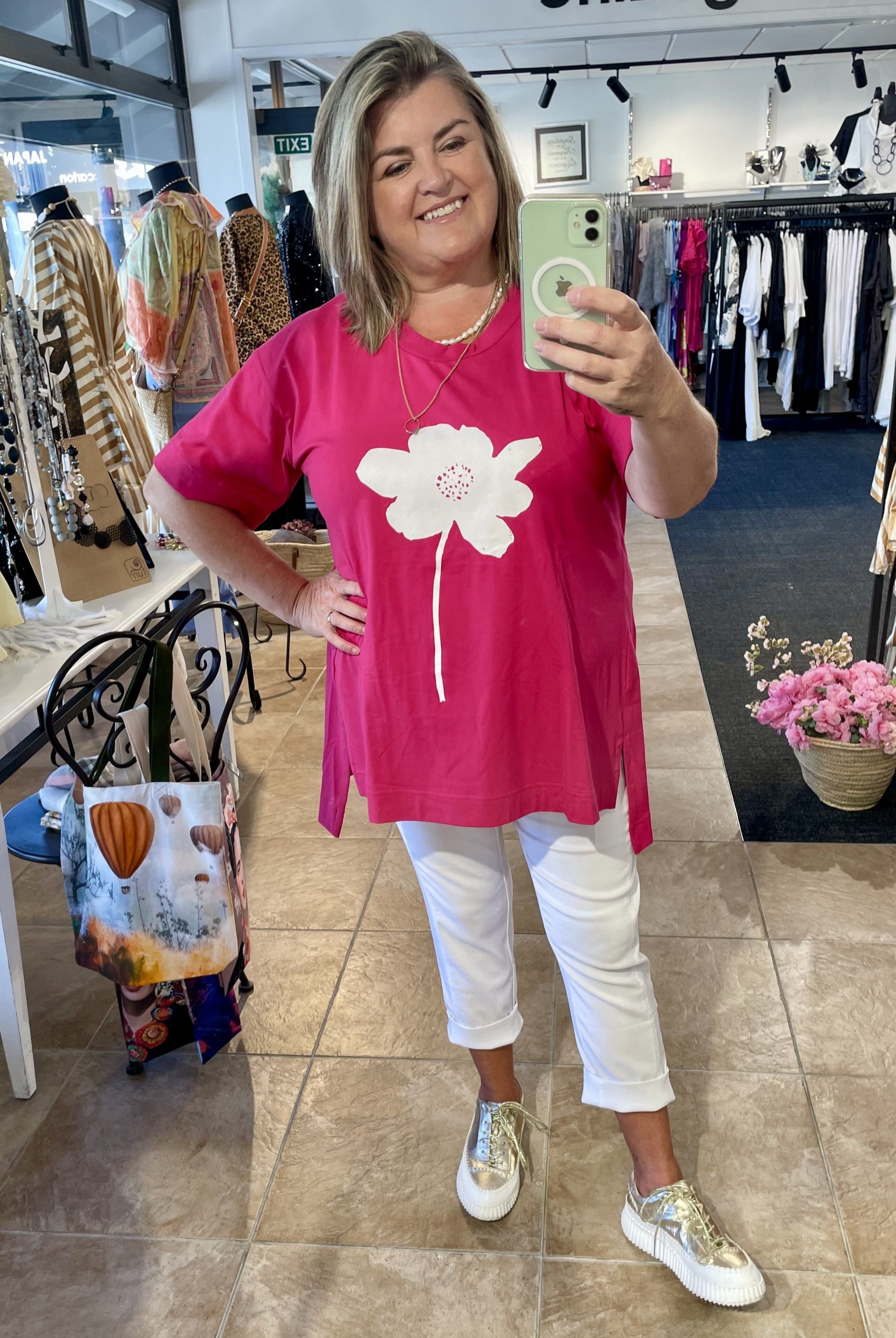 Woman taking a mirror selfie wearing a pink t-shirt with a white flower design in a clothing store.