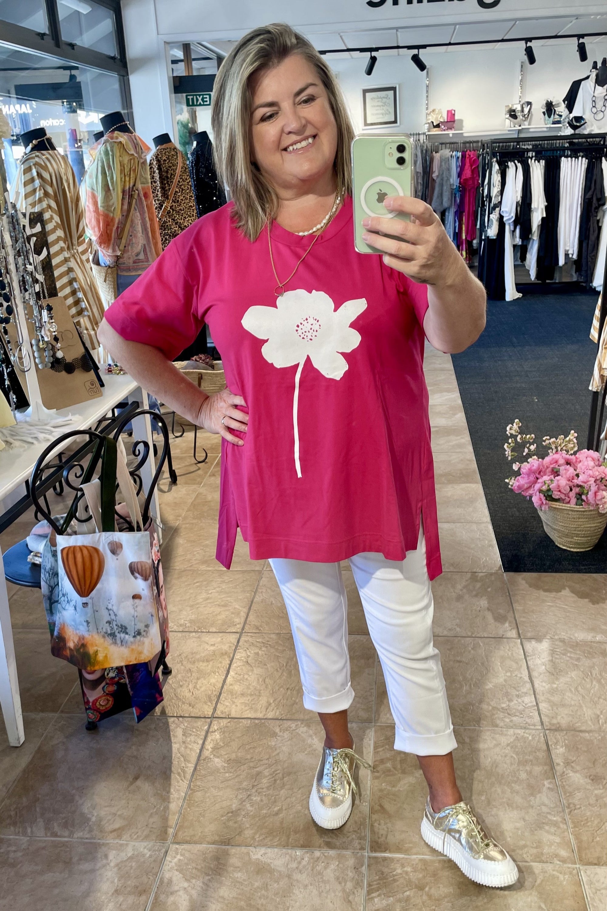 Woman taking a mirror selfie wearing a pink t-shirt with a white flower design in a clothing store.