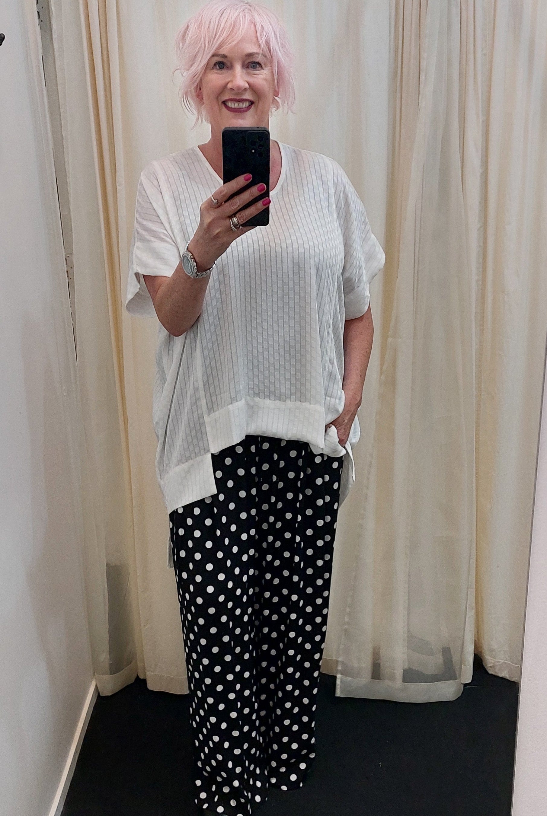 Person with pink hair taking a mirror selfie wearing a white top and black polka dot pants.