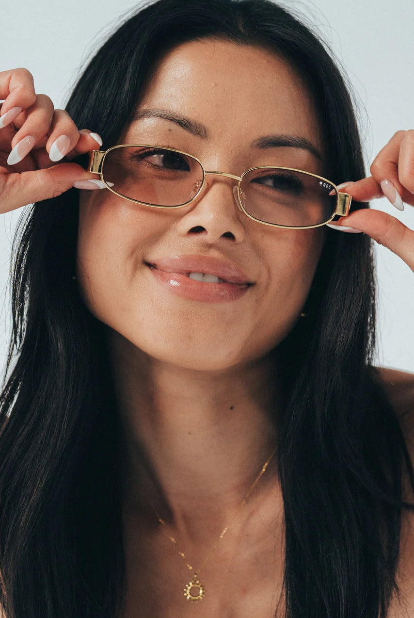 Woman holding gold-framed glasses up to her face against a plain background