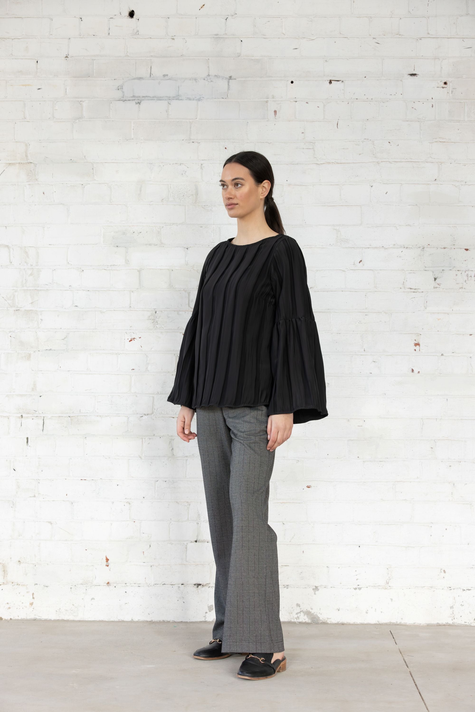 Woman wearing a black top and gray pants standing against a white brick wall.