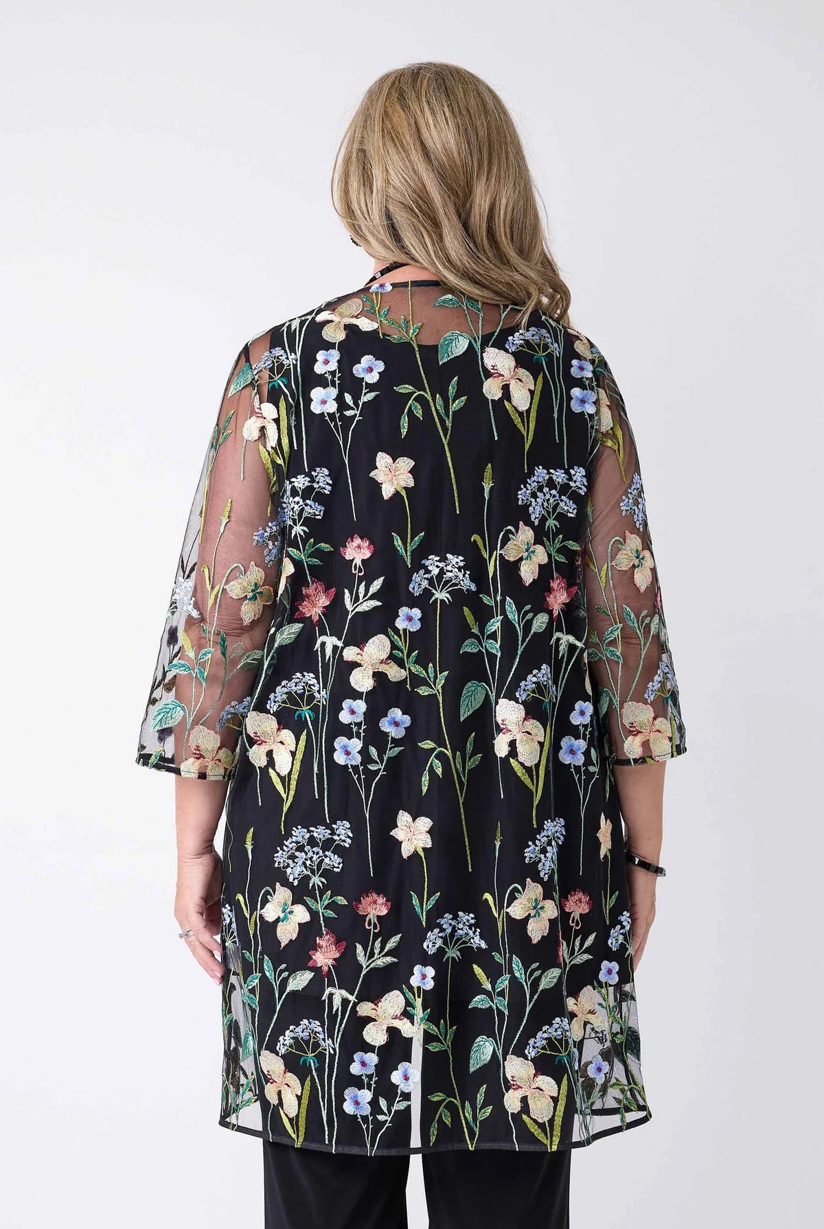 A woman viewed from the back wearing a sheer black tunic-style top with a colorful floral print and elbow-length sleeves, paired with black pants, against a plain light gray studio background.
