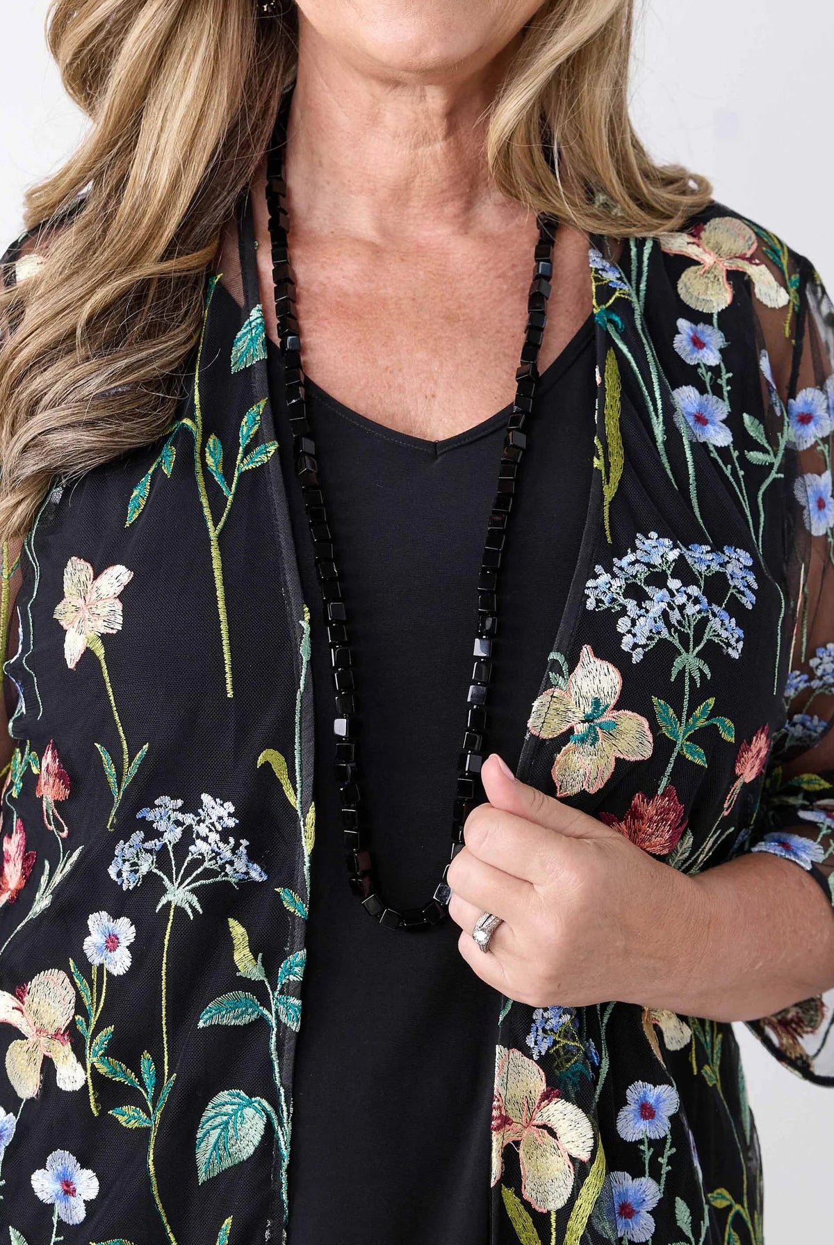 A close-up of a woman wearing a black sheer floral kimono cardigan with embroidered flowers and green leaves over a black top, accessorized with a long black beaded necklace.