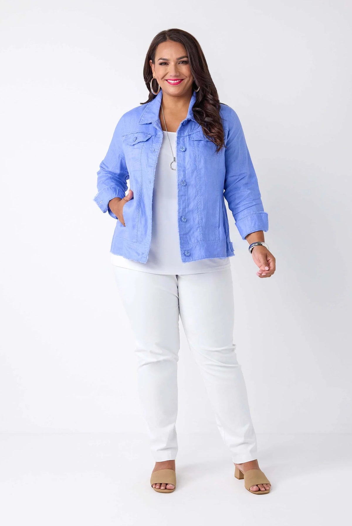 A woman modeling a light blue button-up shirt jacket over a white top and white pants, standing against a plain white studio background.