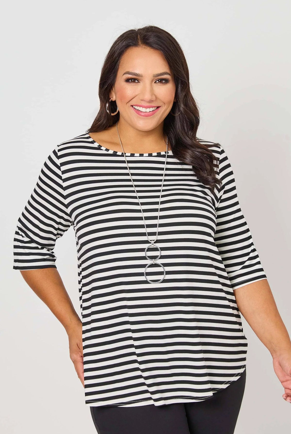 A woman modeling a black and white horizontally striped short-sleeve top with a boat neckline, paired with black pants and a long silver necklace, against a light gray studio background.