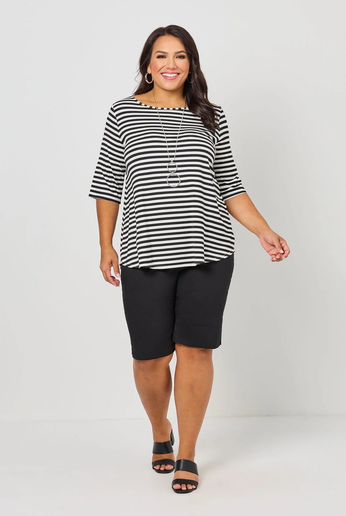 A woman modeling a black and white horizontally striped short-sleeve top with black knee-length shorts and black sandals, standing against a light gray studio background.