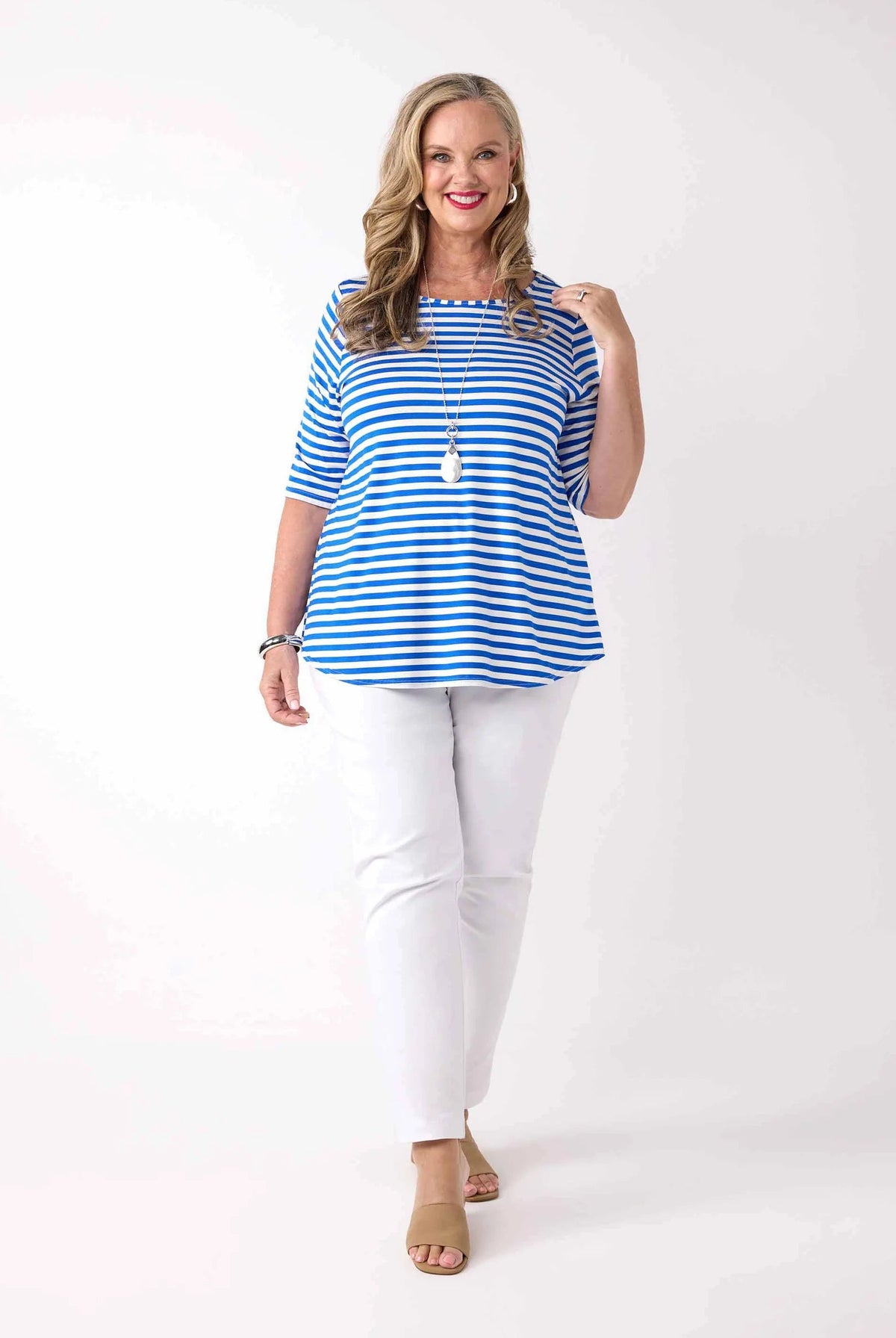 A woman modeling a blue and white striped short-sleeve top with white pants and tan heeled sandals, standing against a plain white studio background.