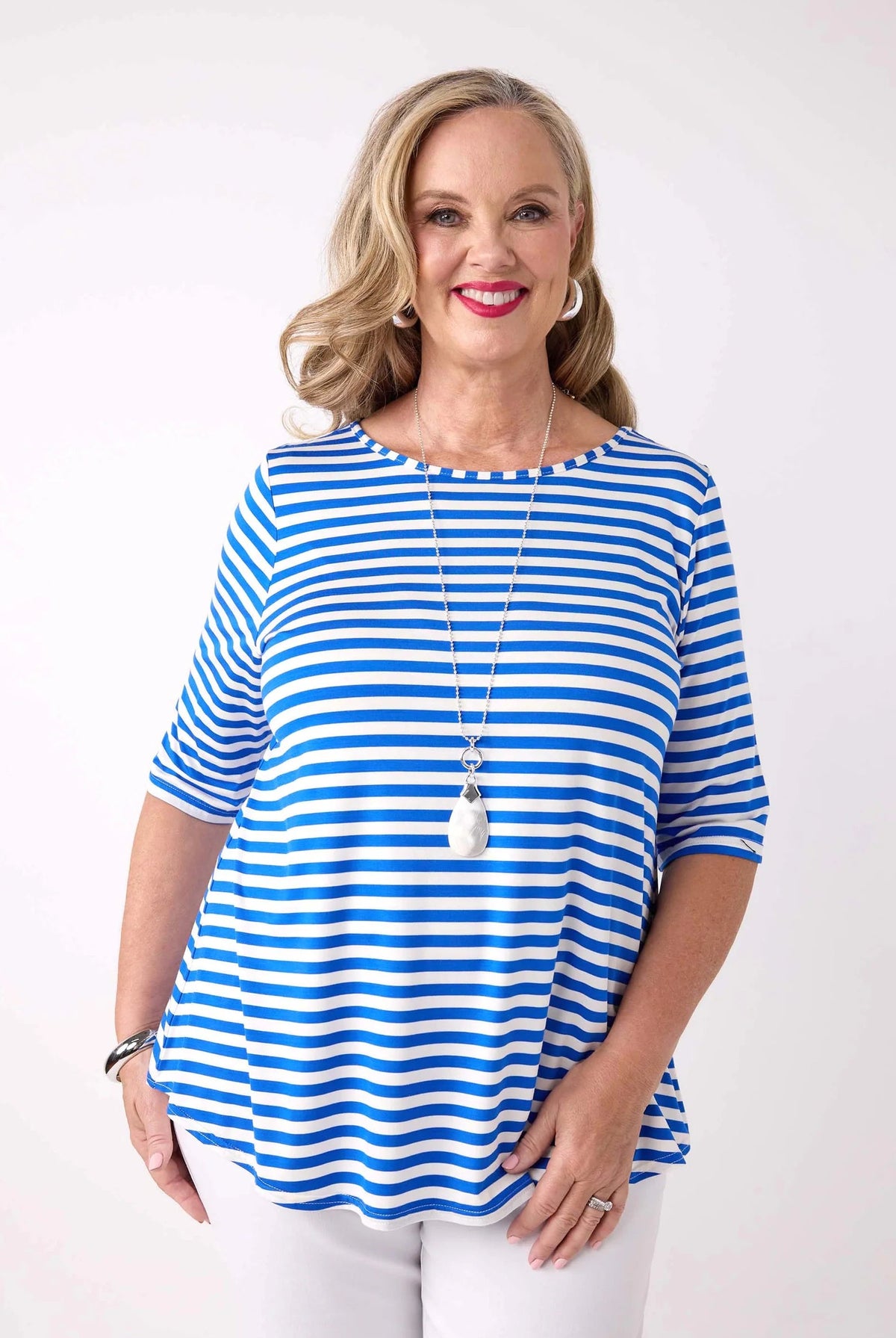 A smiling blonde woman wearing a blue and white horizontally striped short-sleeve top with white pants, standing against a plain white studio background.