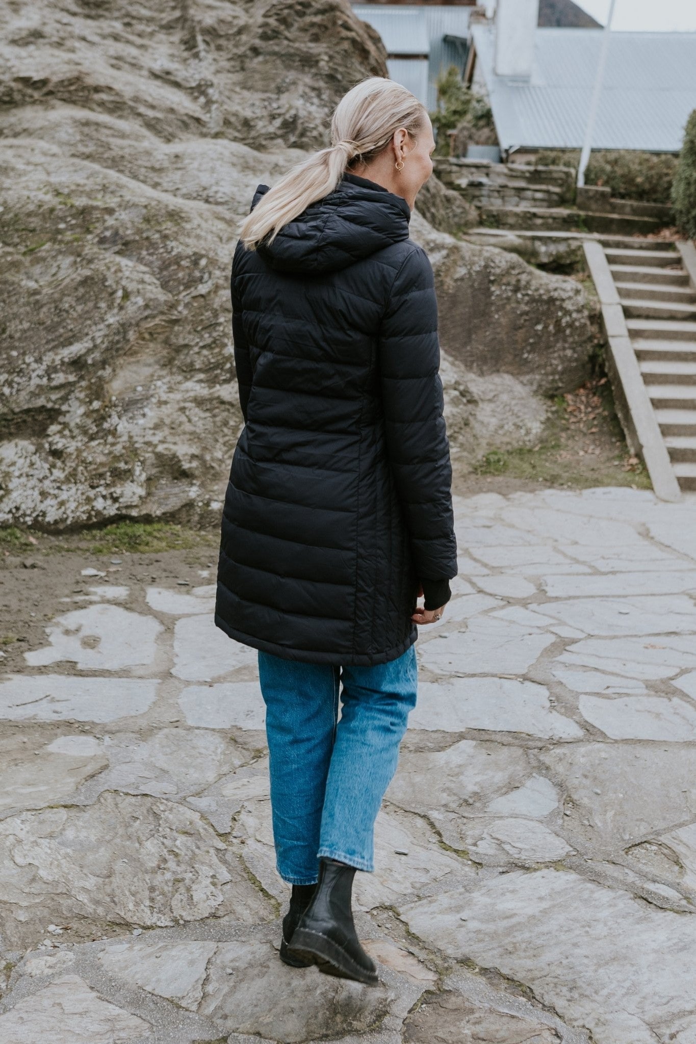 Person wearing a black coat and blue jeans standing on a stone path with large rocks and steps in the background back angle
