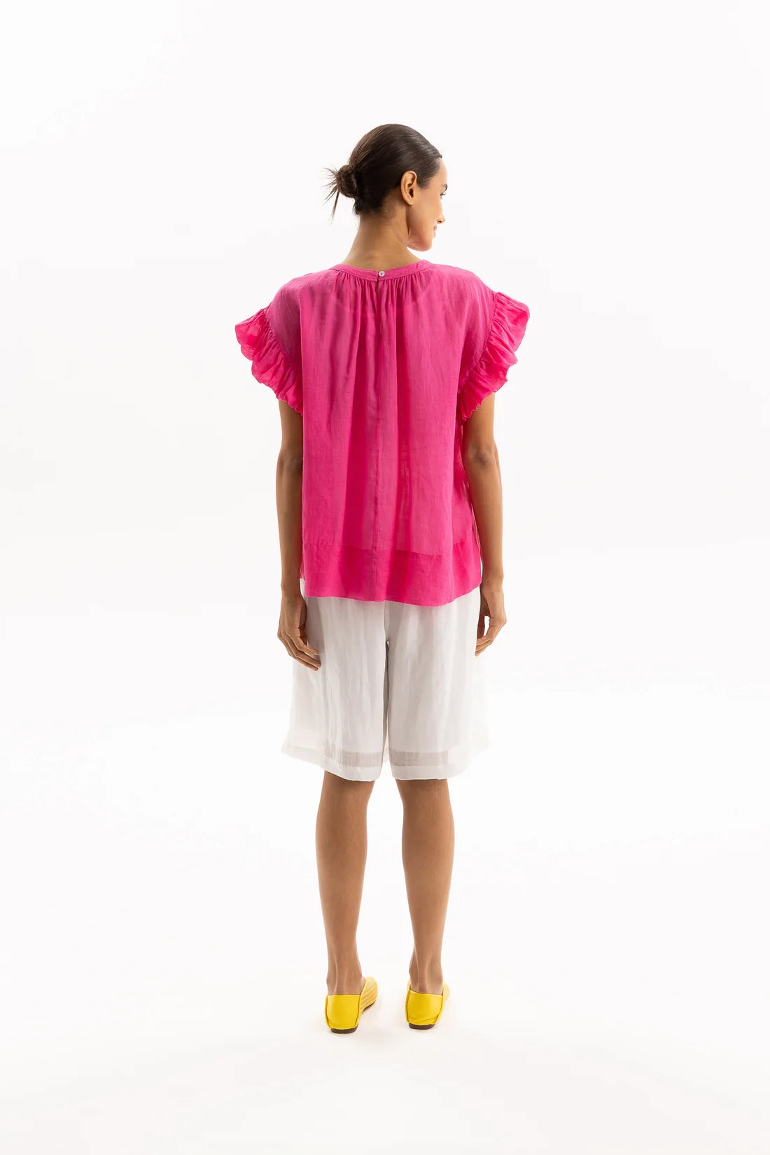 A woman shown from the back wearing a bright pink short-sleeve blouse with ruffled sleeves and white shorts, standing against a plain white studio background.