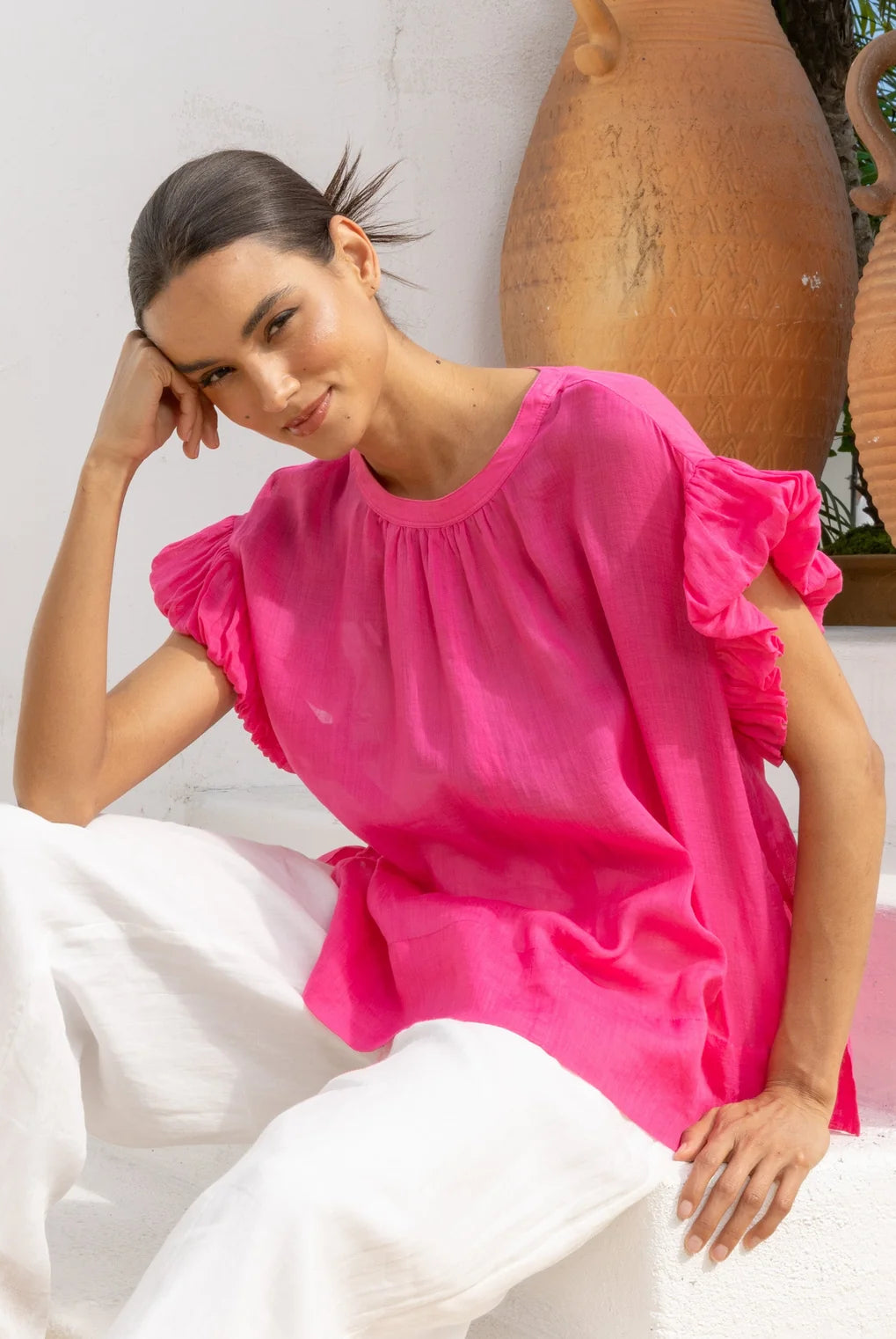 A woman wearing a bright fuchsia pink short-sleeve blouse with ruffled details and white pants, posing in front of a white wall with large terracotta pots.