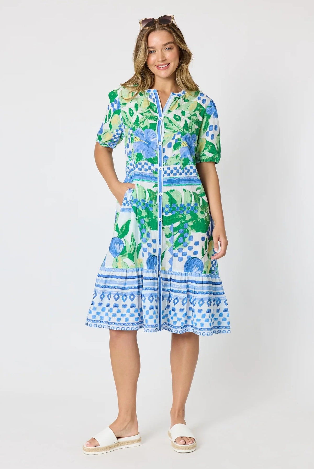 Woman wearing a button up knee length summer dress featuring Mediterranean print with botanical, lemon and blue geometric design against a white background.