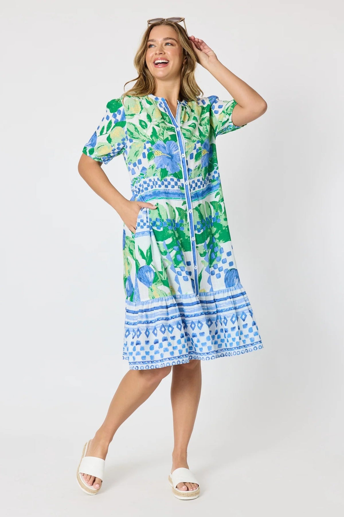Woman wearing a button up knee length summer dress featuring Mediterranean print with botanical, lemon and blue geometric design against a white background.