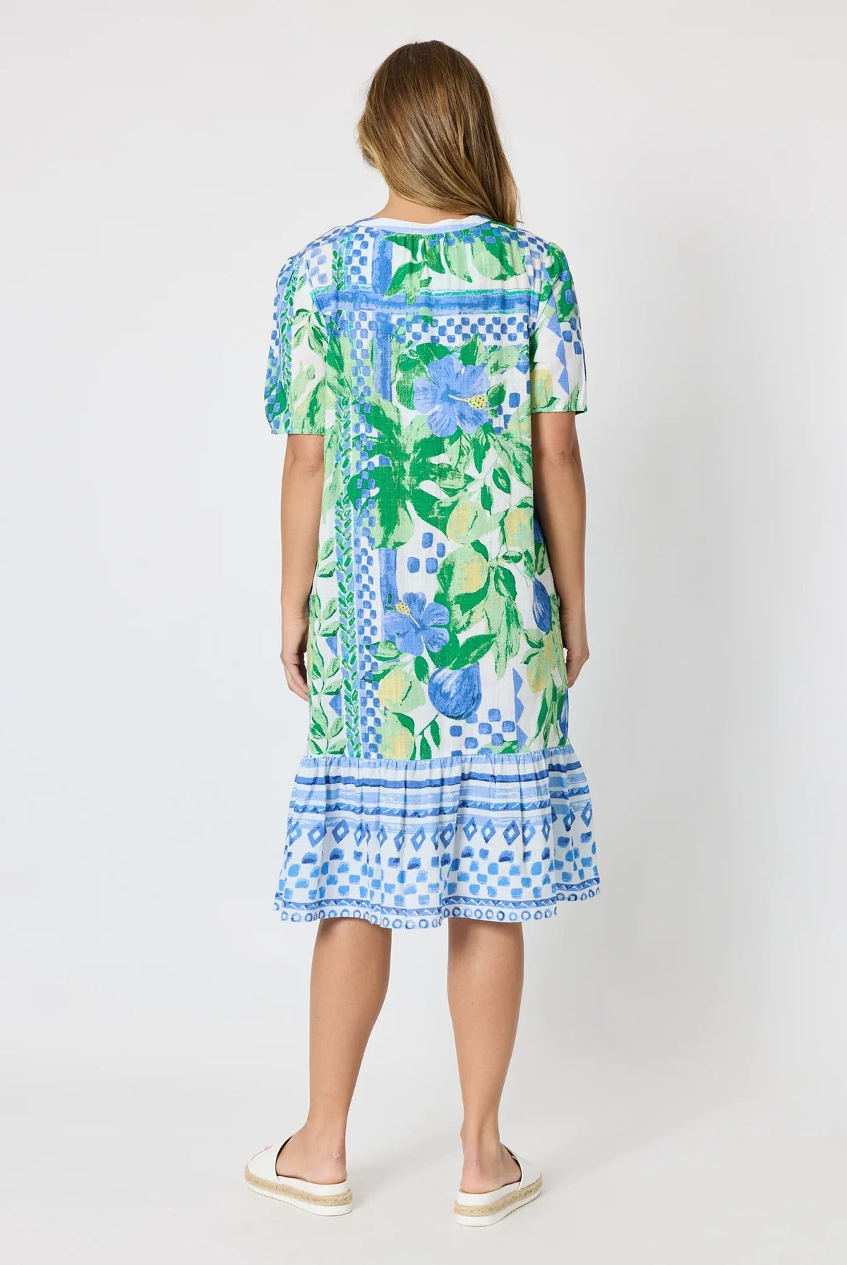 Woman wearing a button up knee length summer dress featuring Mediterranean print with botanical, lemon and blue geometric design against a white background.